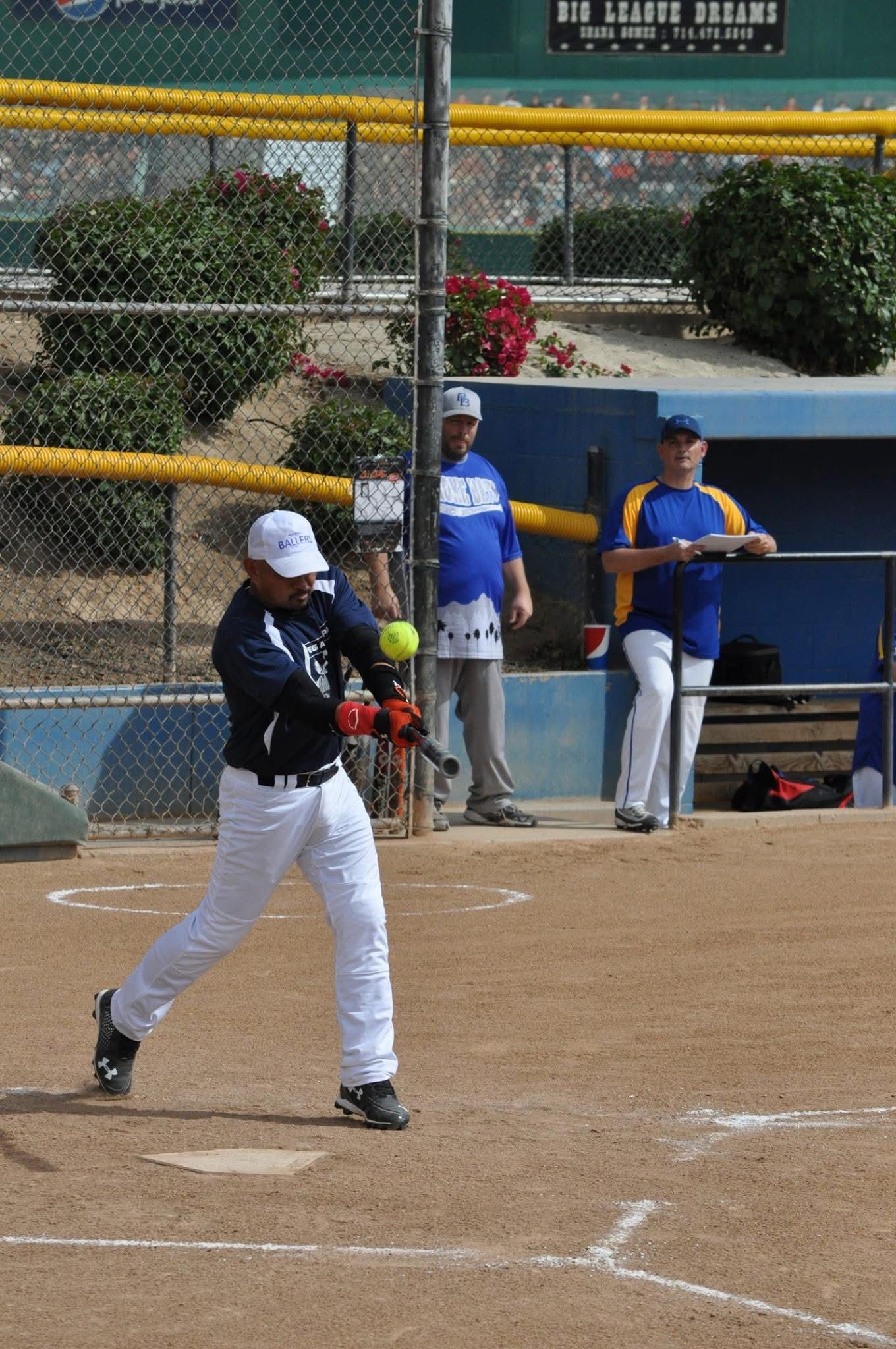 Softball batter in black swings at a pitch on a dirt field, with players and a dugout in the background