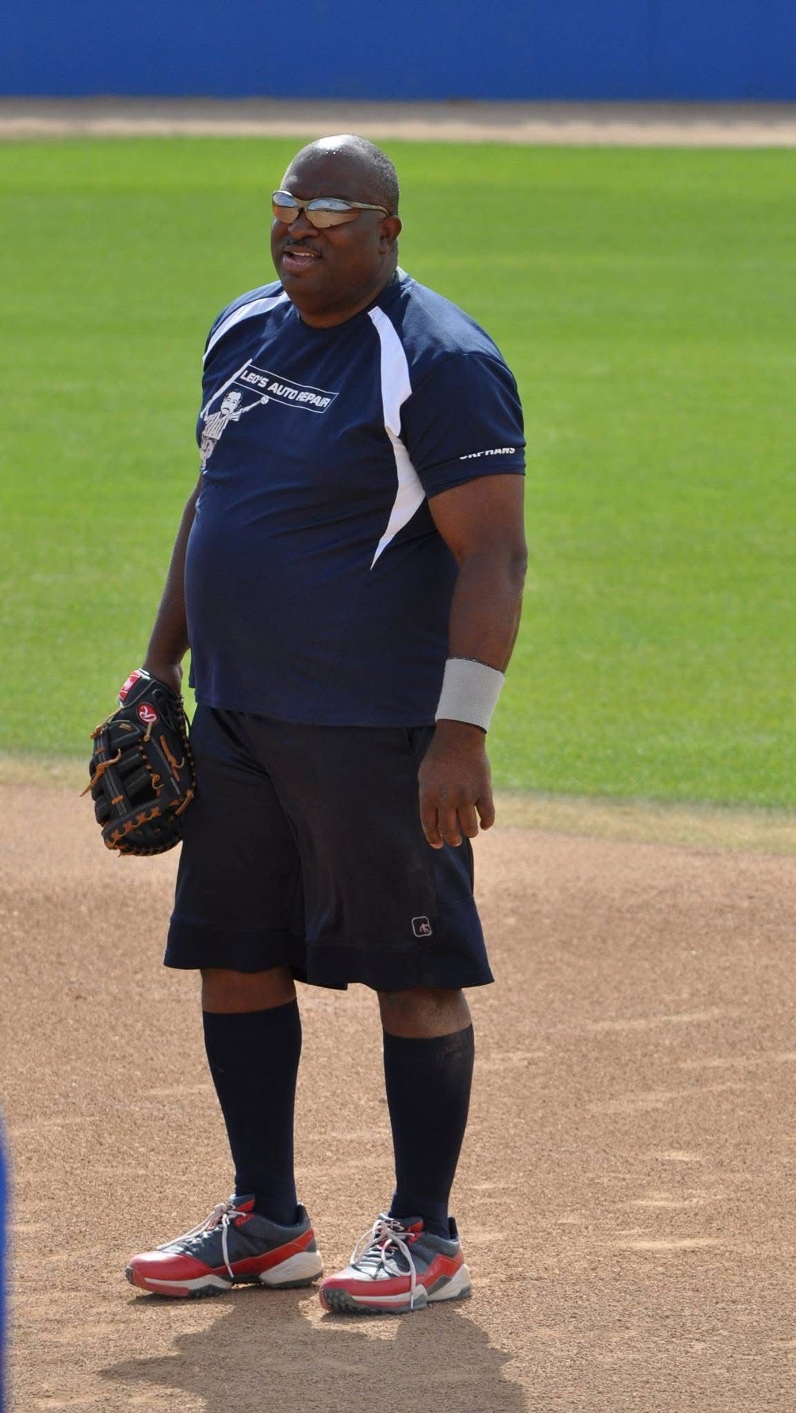 Person standing on a baseball field holding a glove, wearing a navy shirt, shorts, and red cleats.