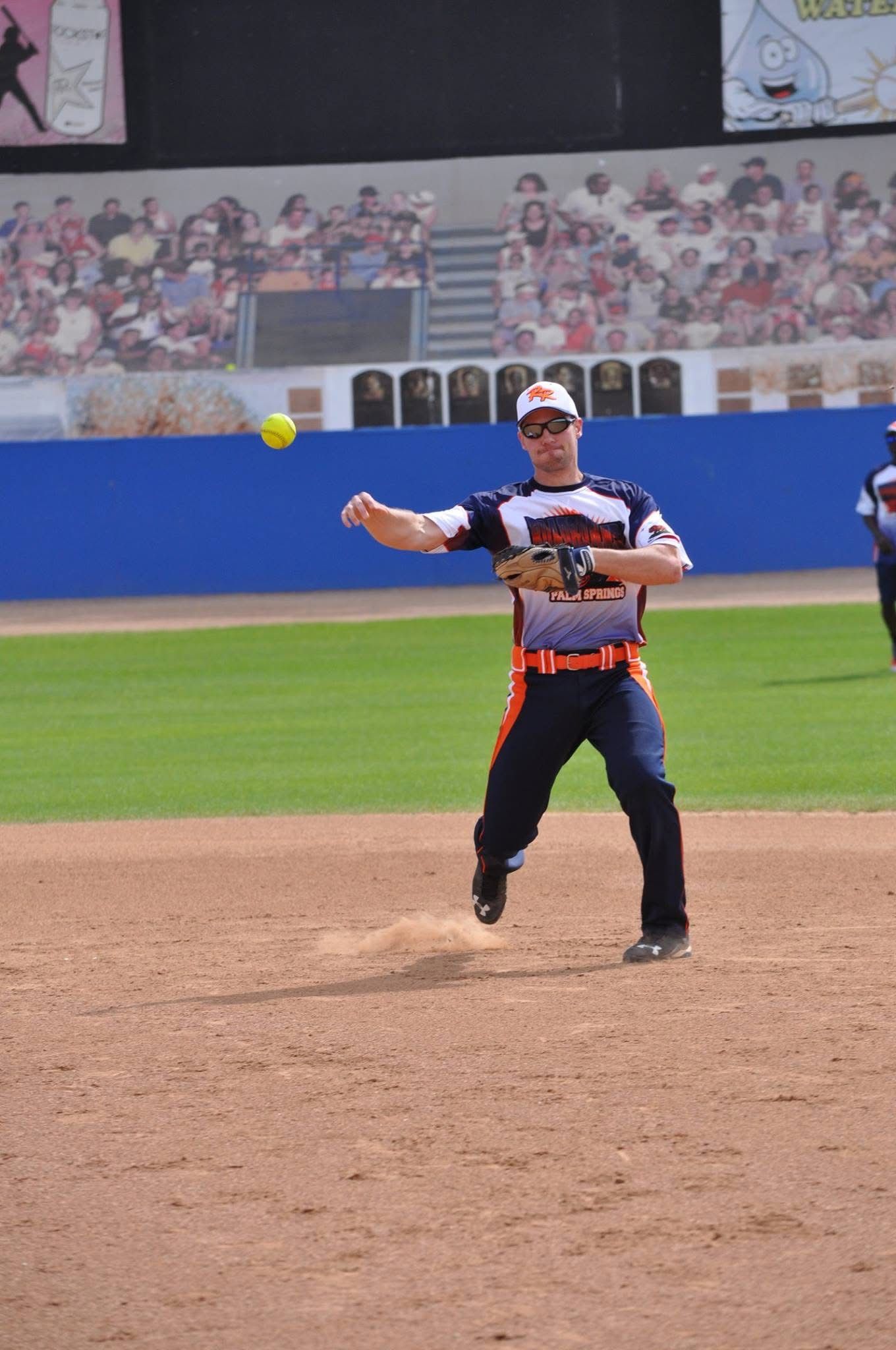 Softball pitcher in black uniform mid-throw on the mound, with a ball in motion and stadium crowd behind.
