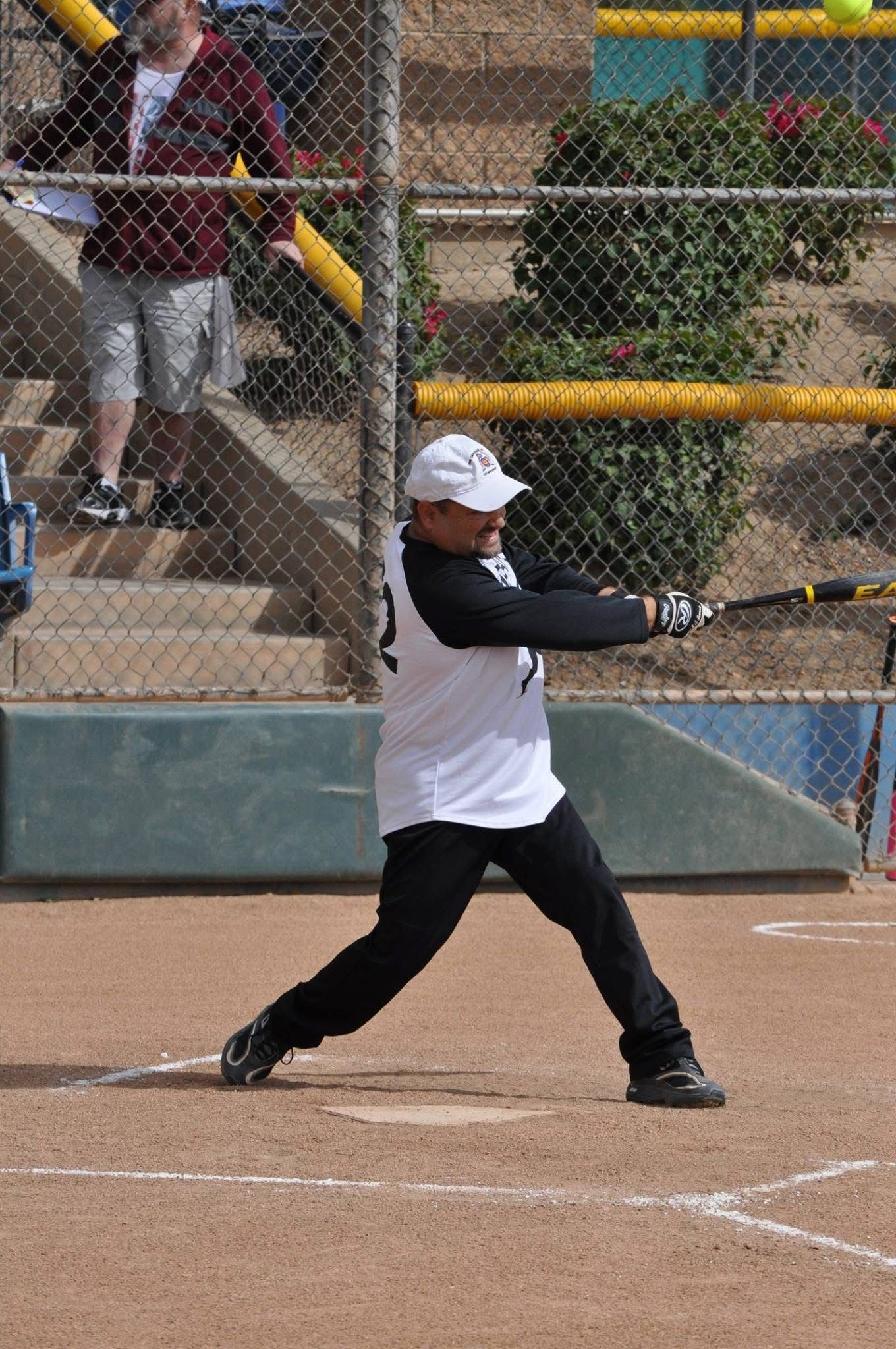 Baseball batter in white and black swings at a pitch on a dirt field near a chain-link fence.