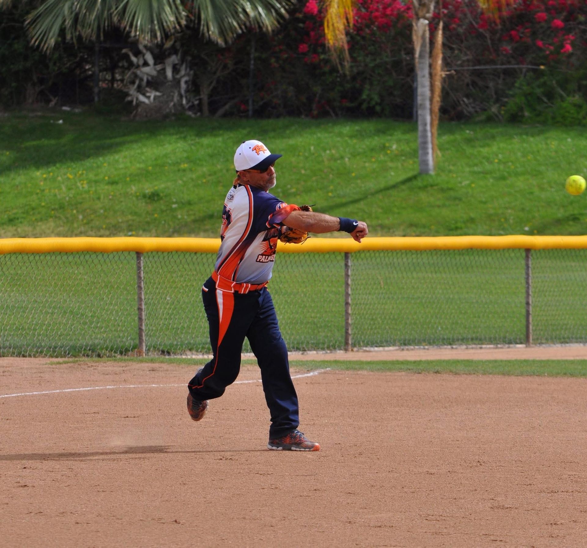 Softball player in gray and orange swings a bat on a dirt field near an outfield fence.