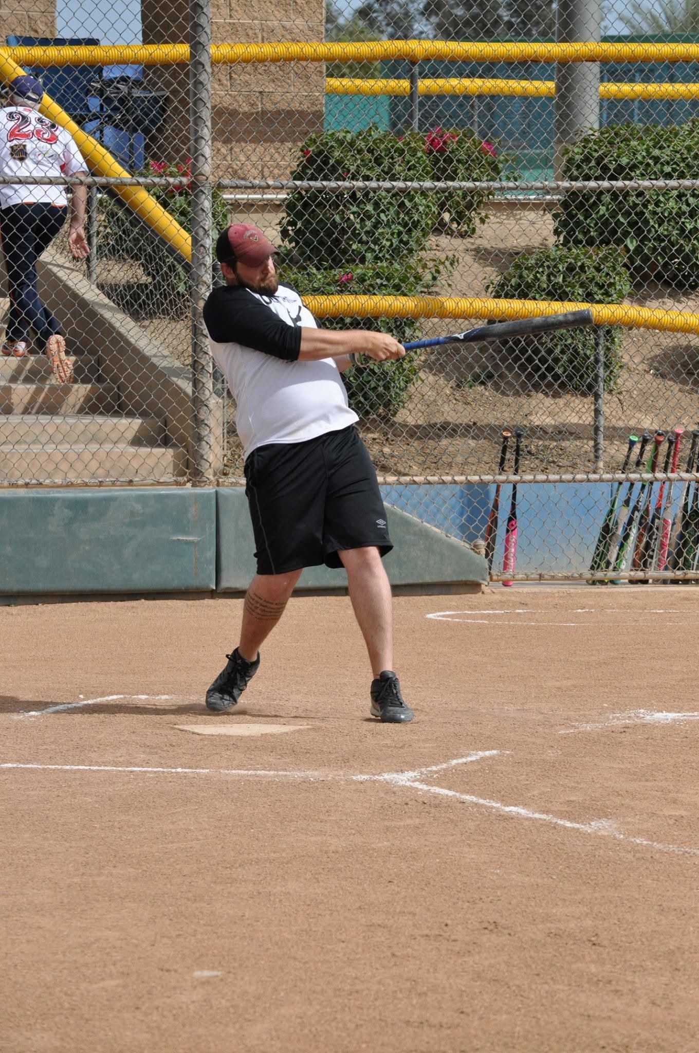 Baseball batter swinging at a pitch on a dirt field, with bleachers and a fence in the background