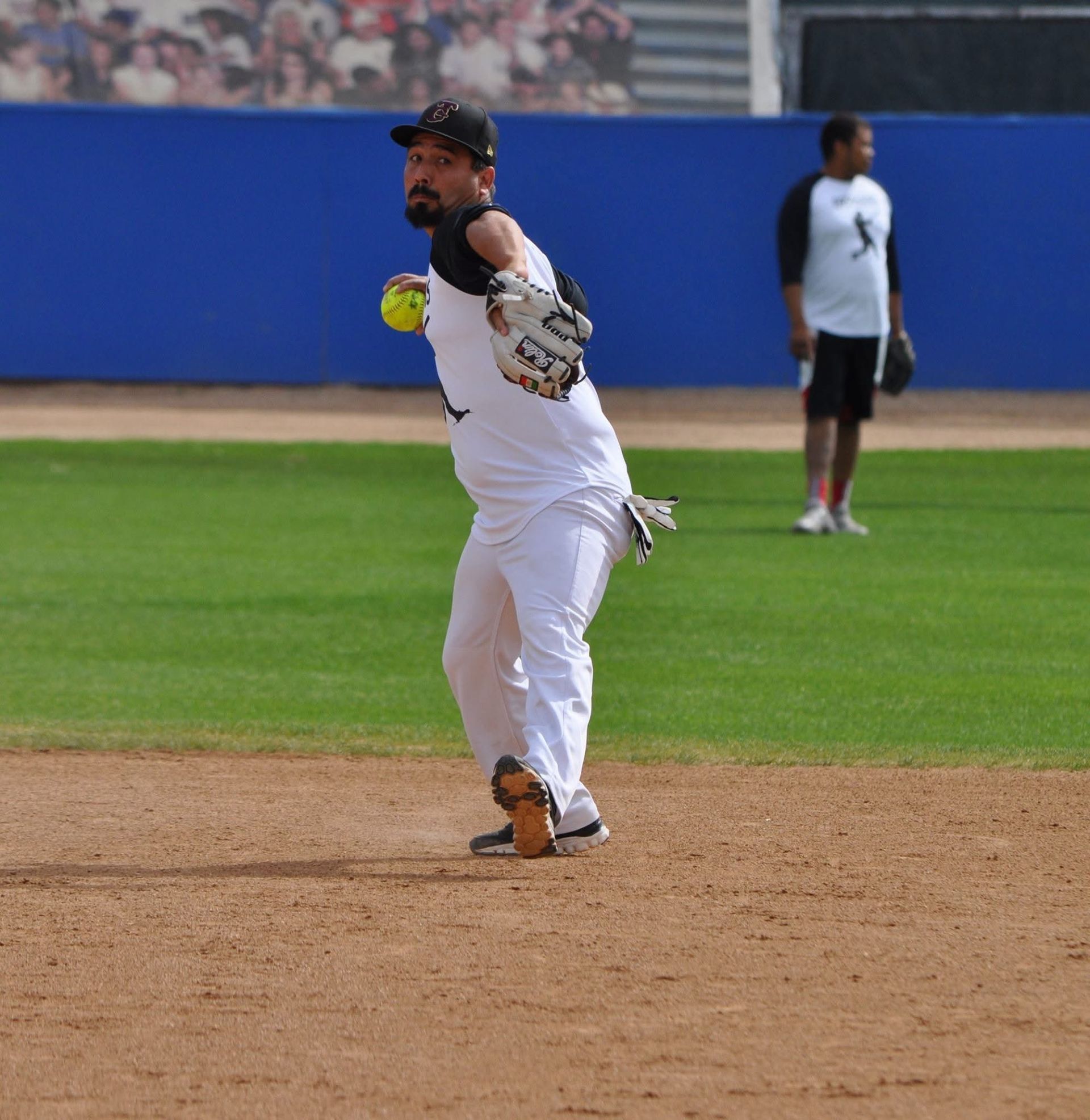 Baseball player in white uniform throws a ball on the infield with outfield wall behind him.