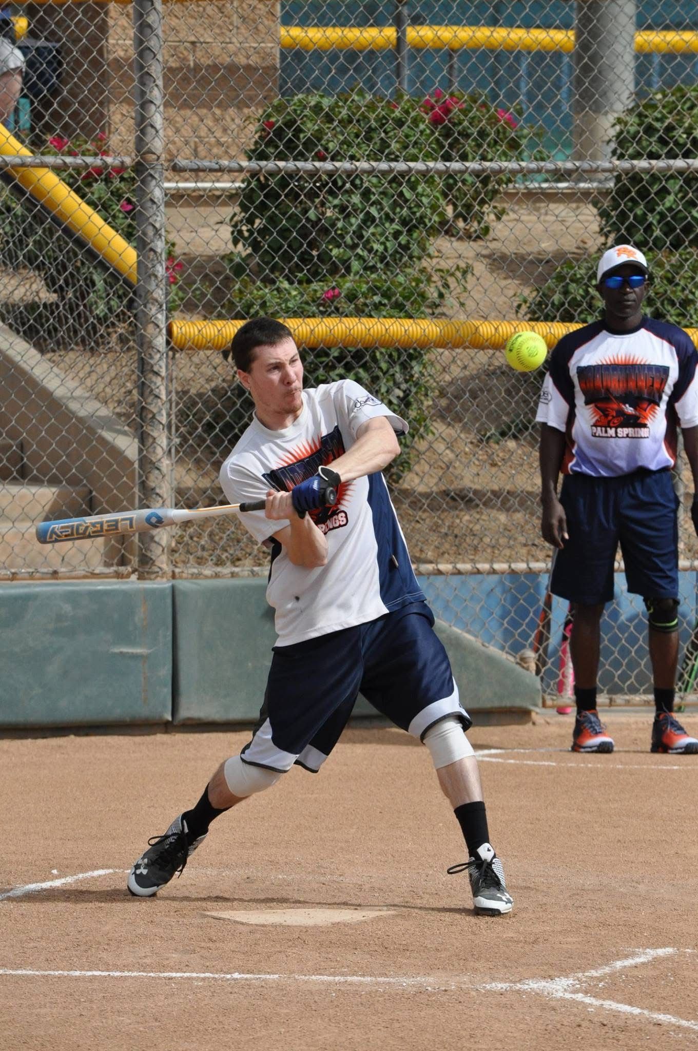 Softball player in white swings at a pitch on a dirt field, with a teammate standing behind the fence.