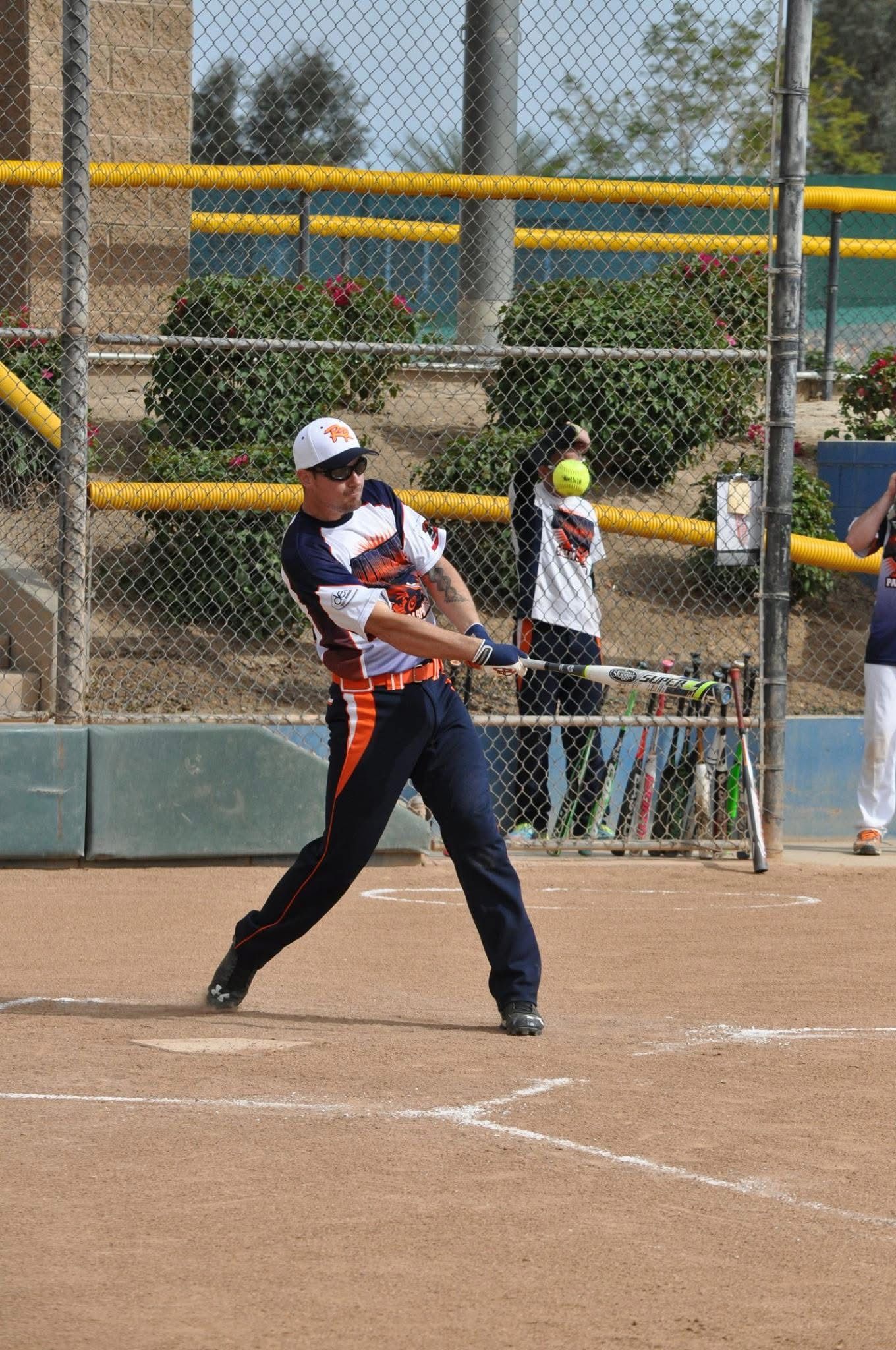 Softball batter swinging at home plate on a dirt field, with fence and spectators in the background