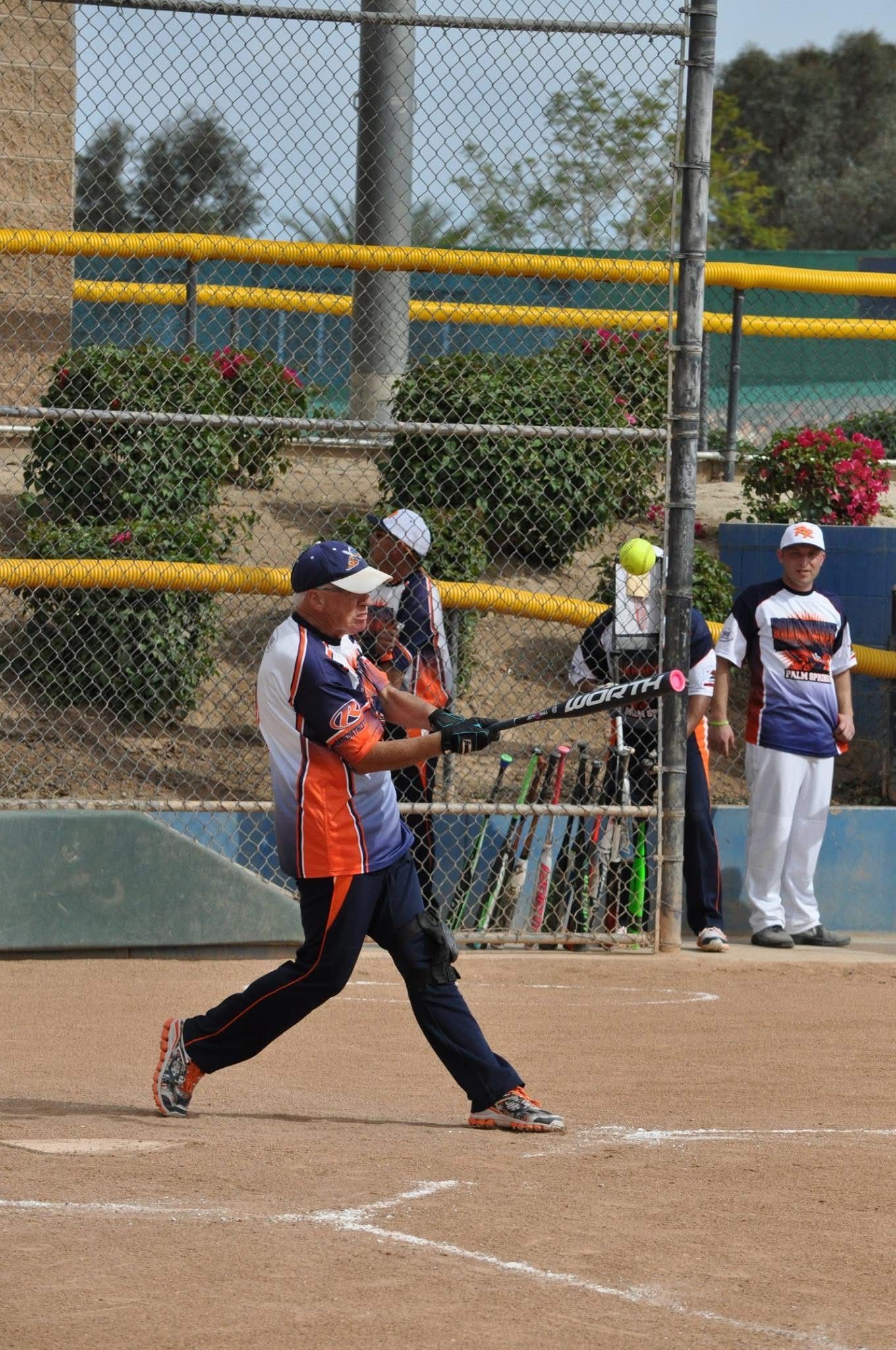 Softball player in orange jersey swings bat at home plate on a dirt field, with spectators and fence behind.