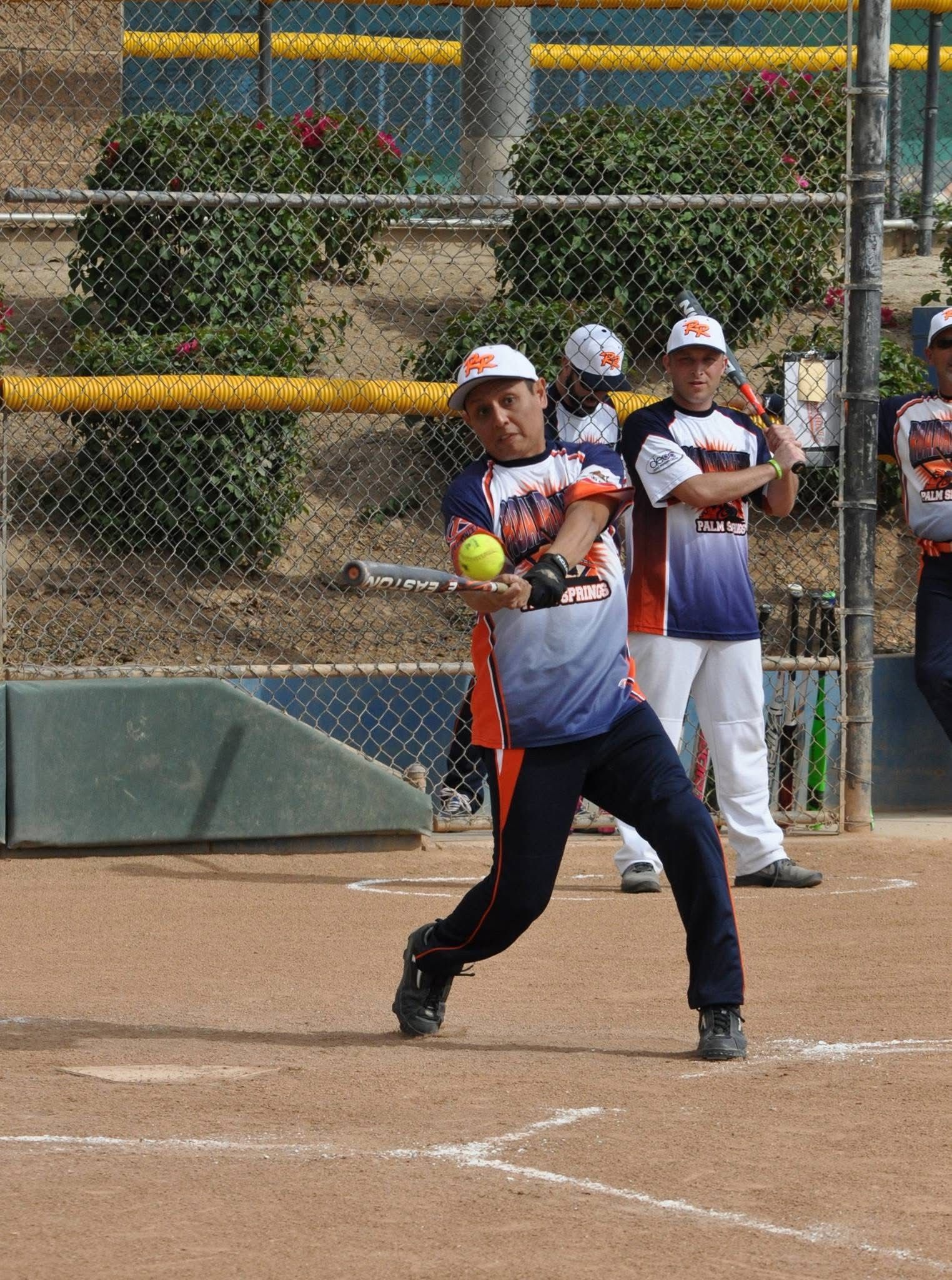 Softball batter in gray and red swings at a pitch on a dirt field, with teammates watching behind her.