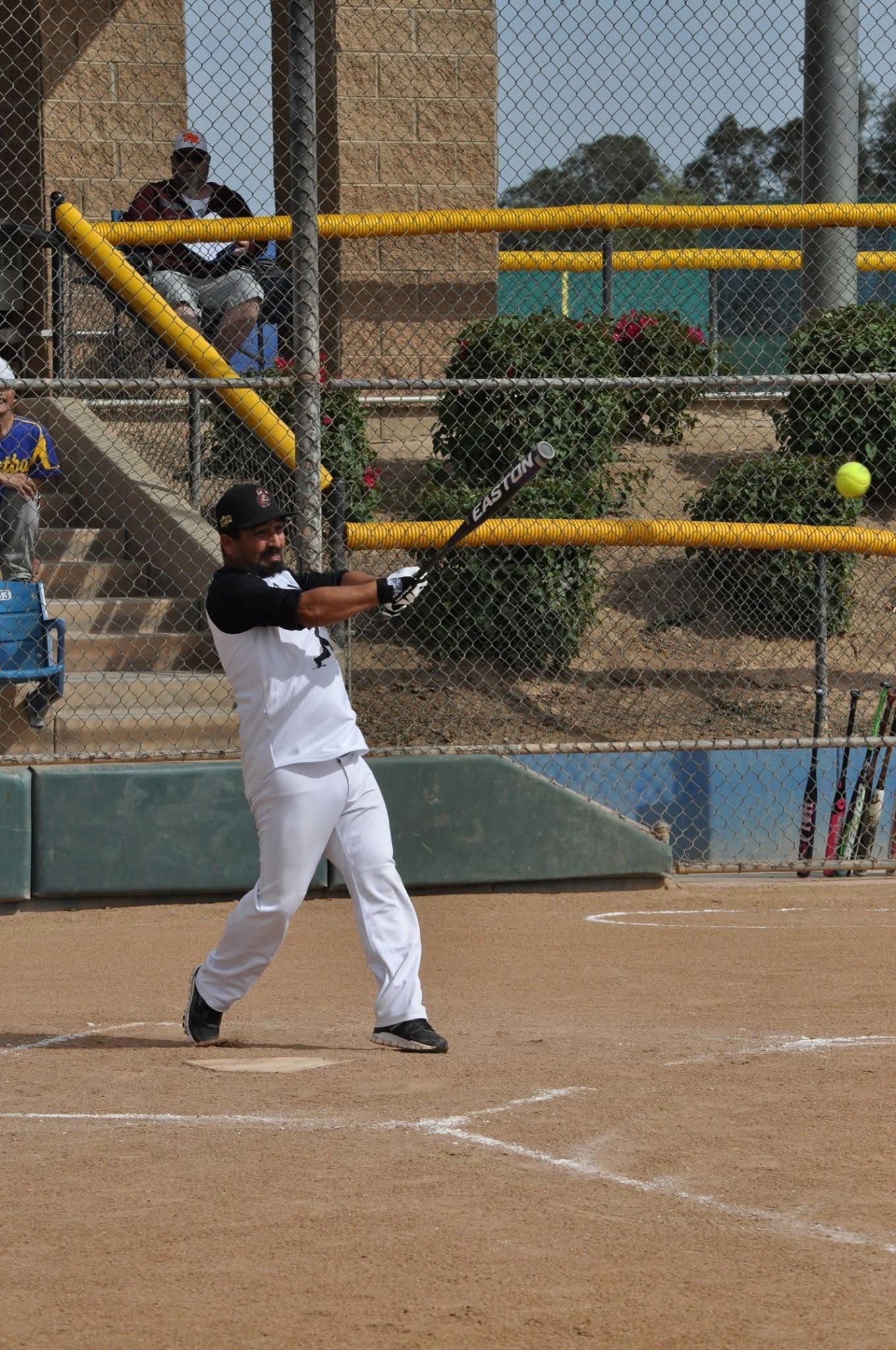 Baseball batter in white uniform swings a bat on a dirt field near the fence.