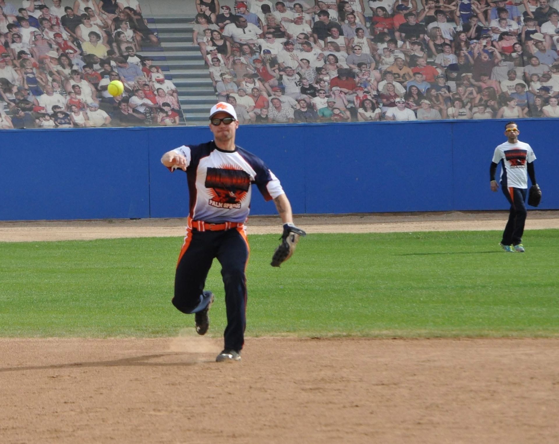 Baseball player in white and navy throws a ball on the field, with another player in the background.