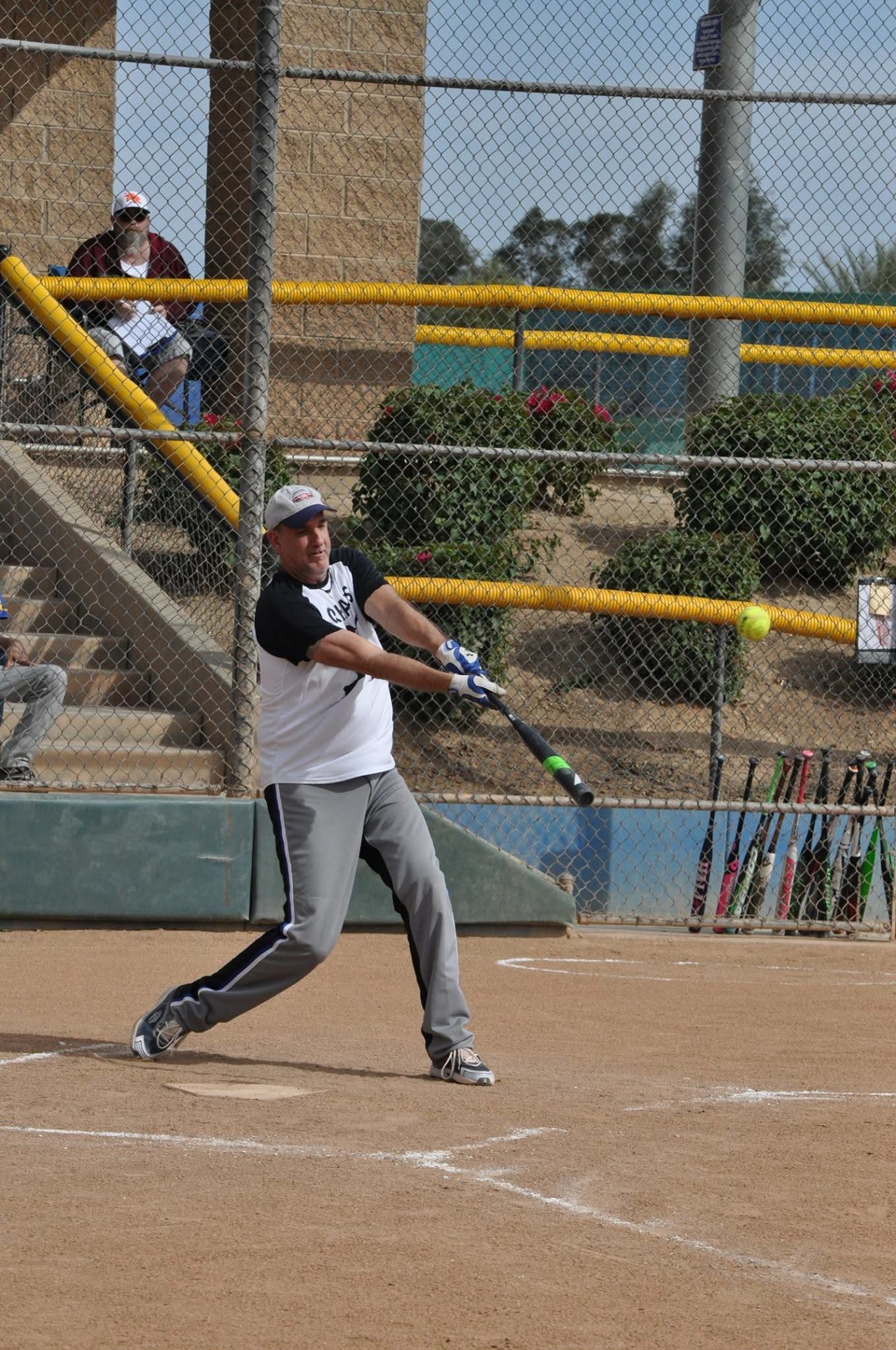 Softball player swinging a bat at home plate on a dirt field, with yellow railings in the background