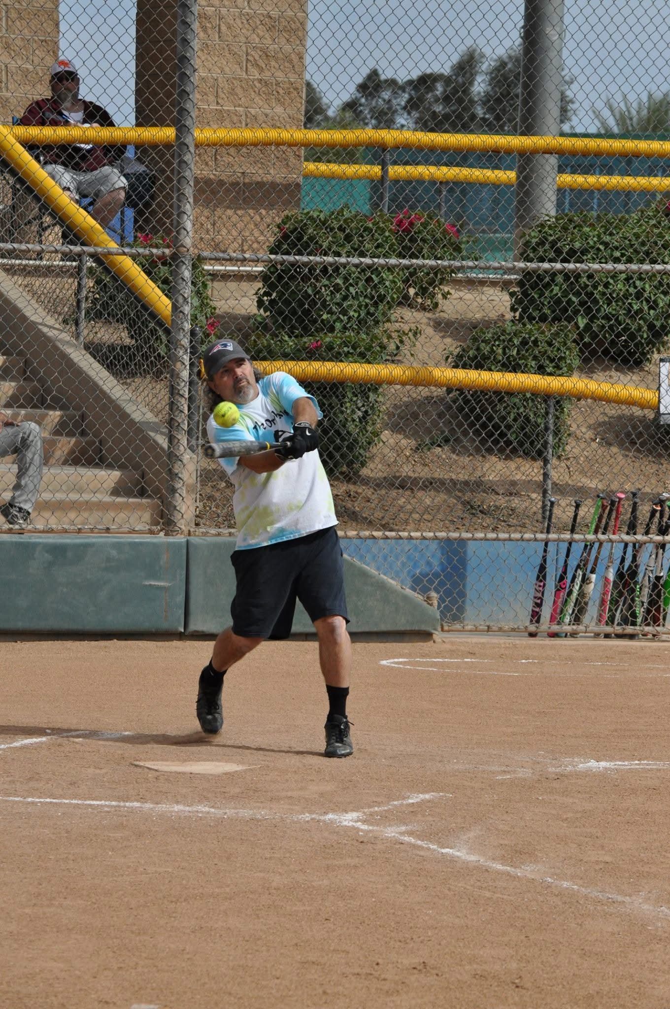Softball batter mid-swing at home plate on a dirt field, with a chain-link fence and yellow railings behind.