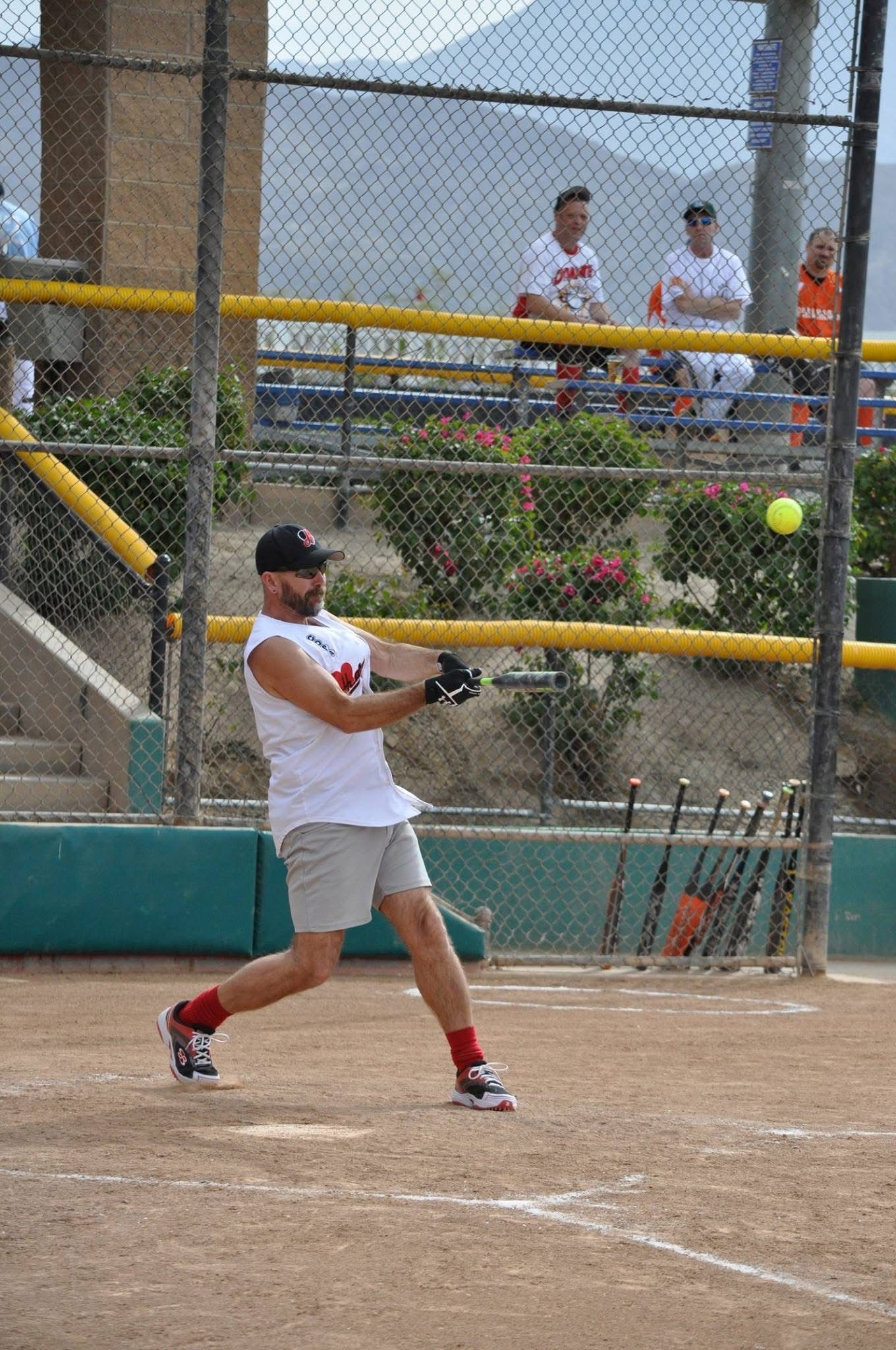 Baseball player in white swings at a ball on a dirt field, with spectators behind a chain-link fence.