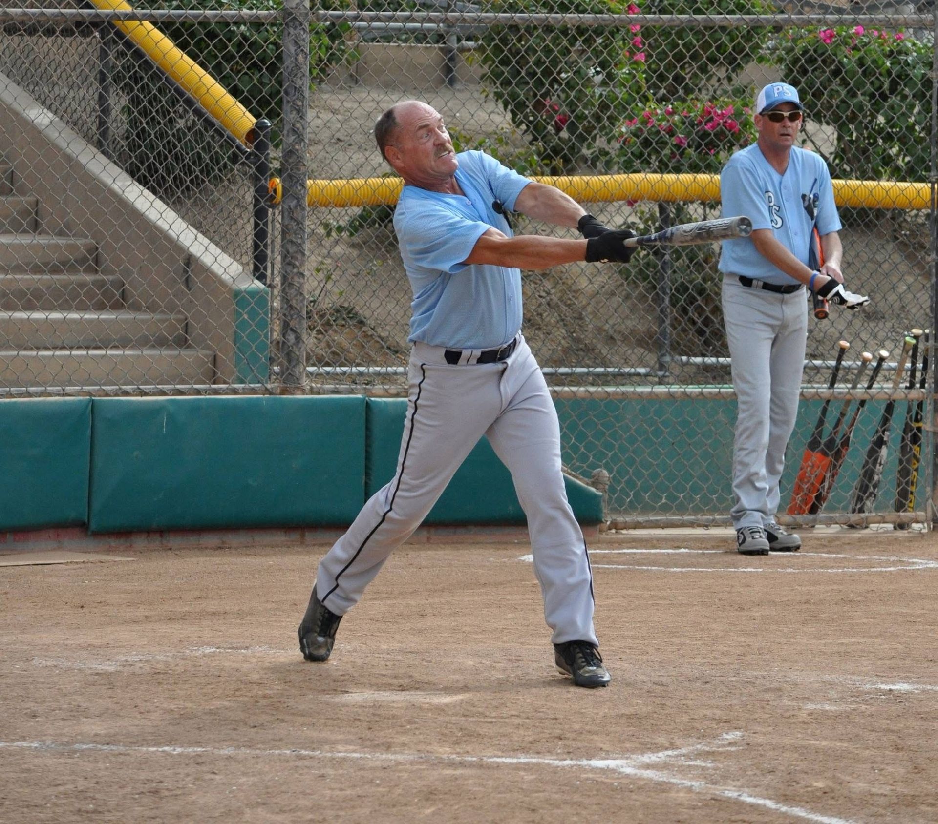 Baseball player in gray uniform swings at a pitch on a dirt field, teammate standing behind him