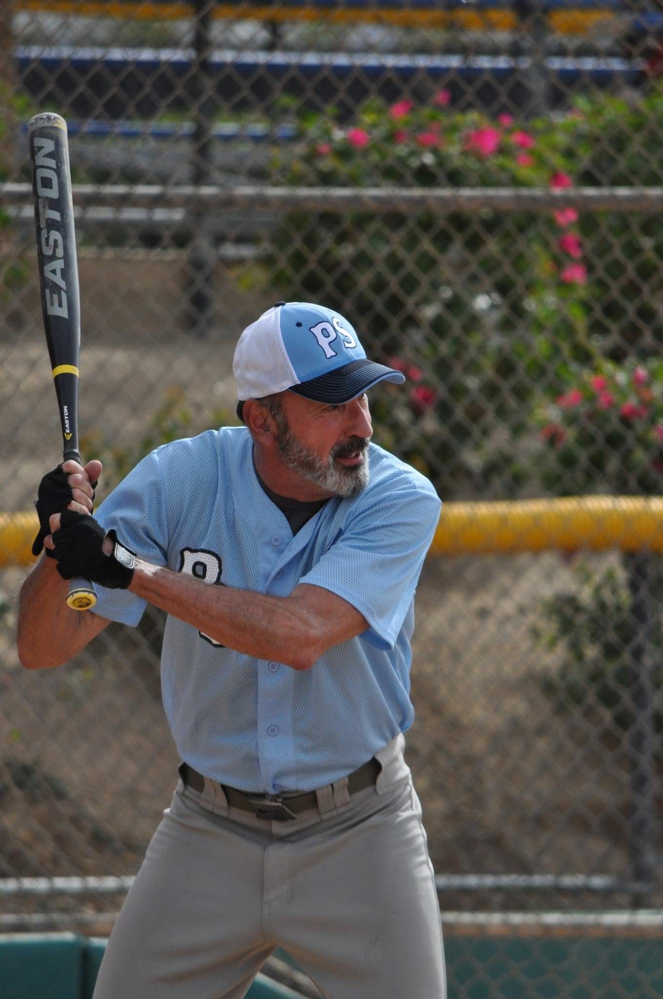 Baseball batter in blue jersey and cap at the plate, holding a bat near a chain-link fence.