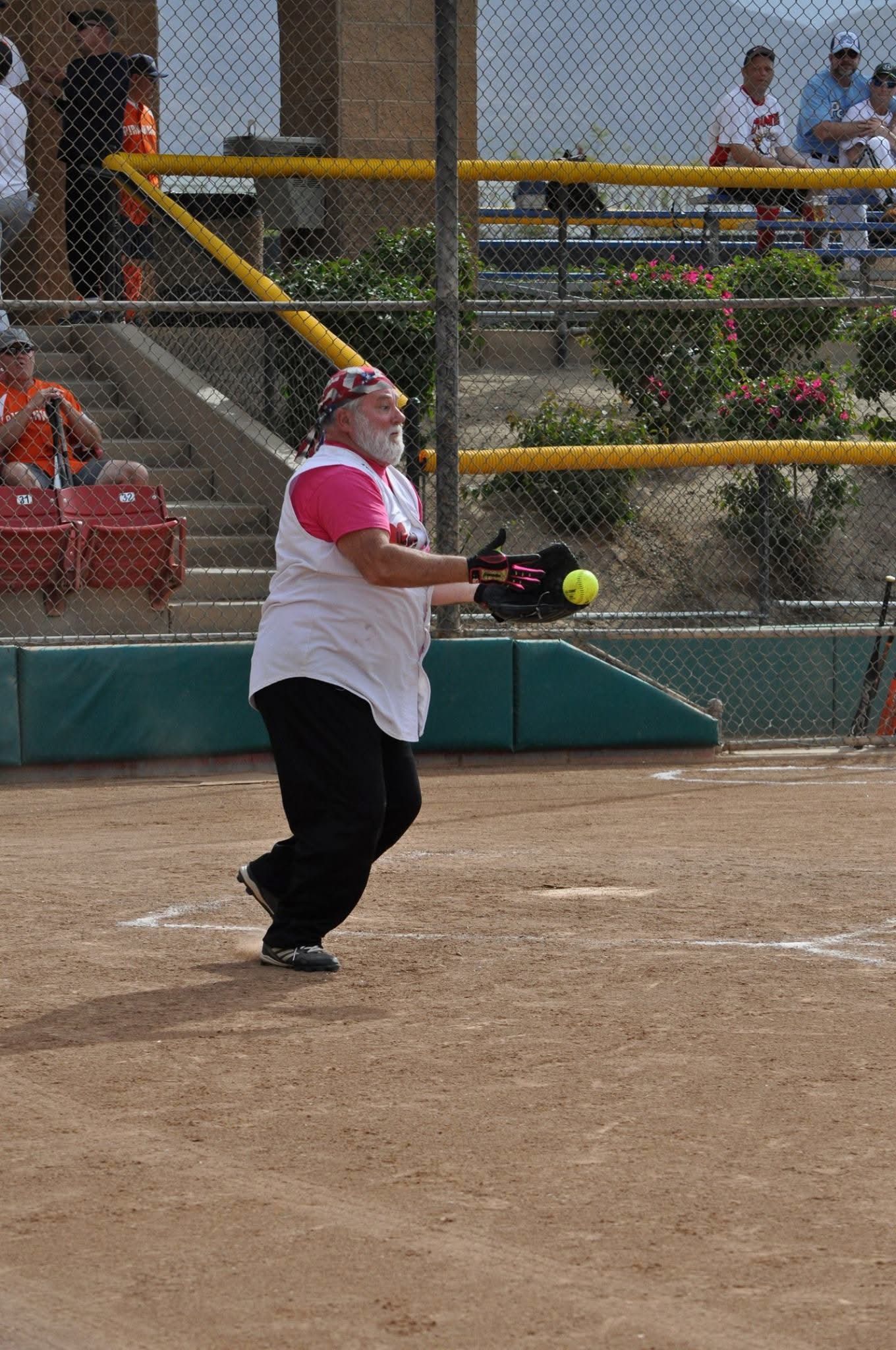 Softball player in white and pink swings at a yellow ball on a dirt field during a game.
