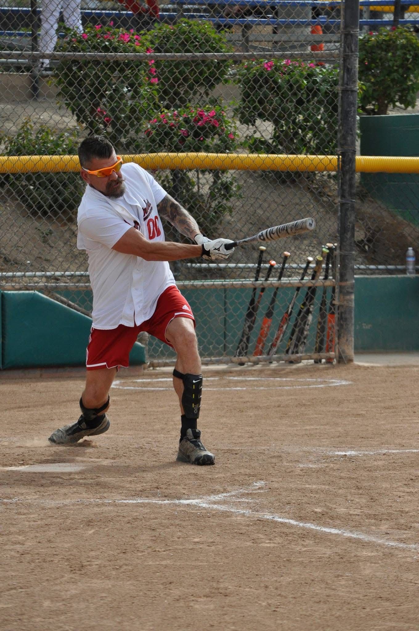 Baseball player in white and red swings a bat on a dirt field, with a chain-link fence behind him.
