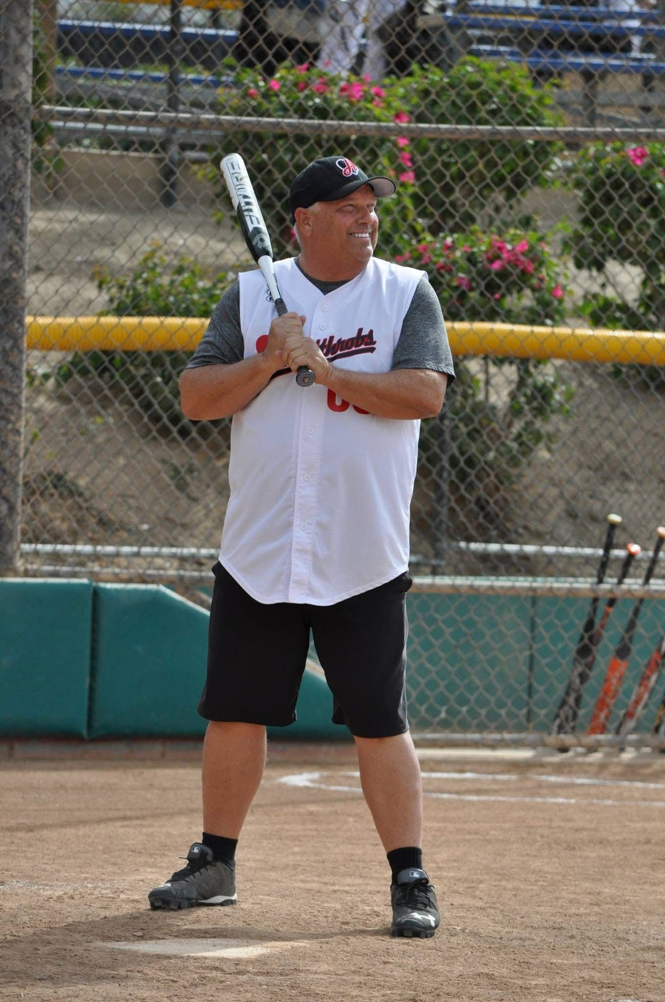 Baseball player in white jersey and black shorts stands ready at bat on a dirt field.