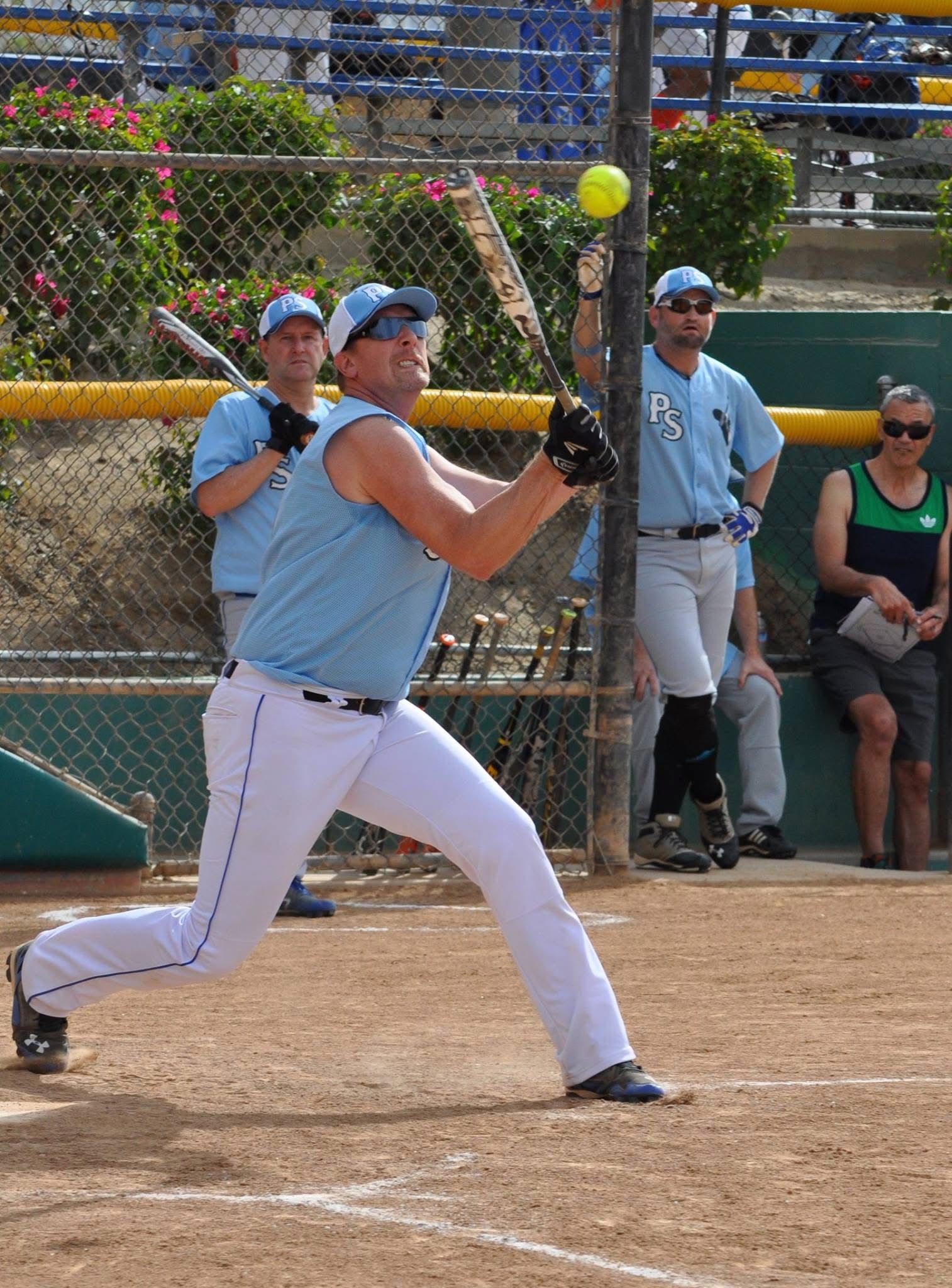 Softball pitcher in light blue uniform throws a pitch on a dirt field with spectators behind a chain-link fence
