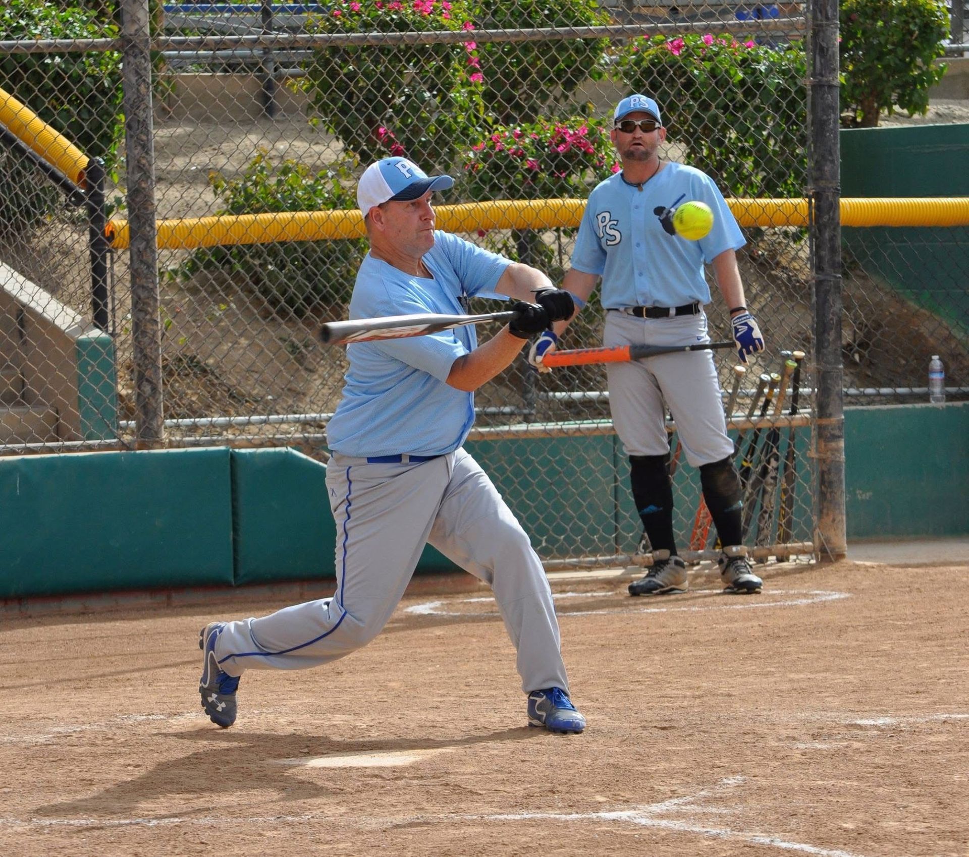 Baseball player in light blue uniform swinging a bat on a dirt field, with teammate behind him near the fence