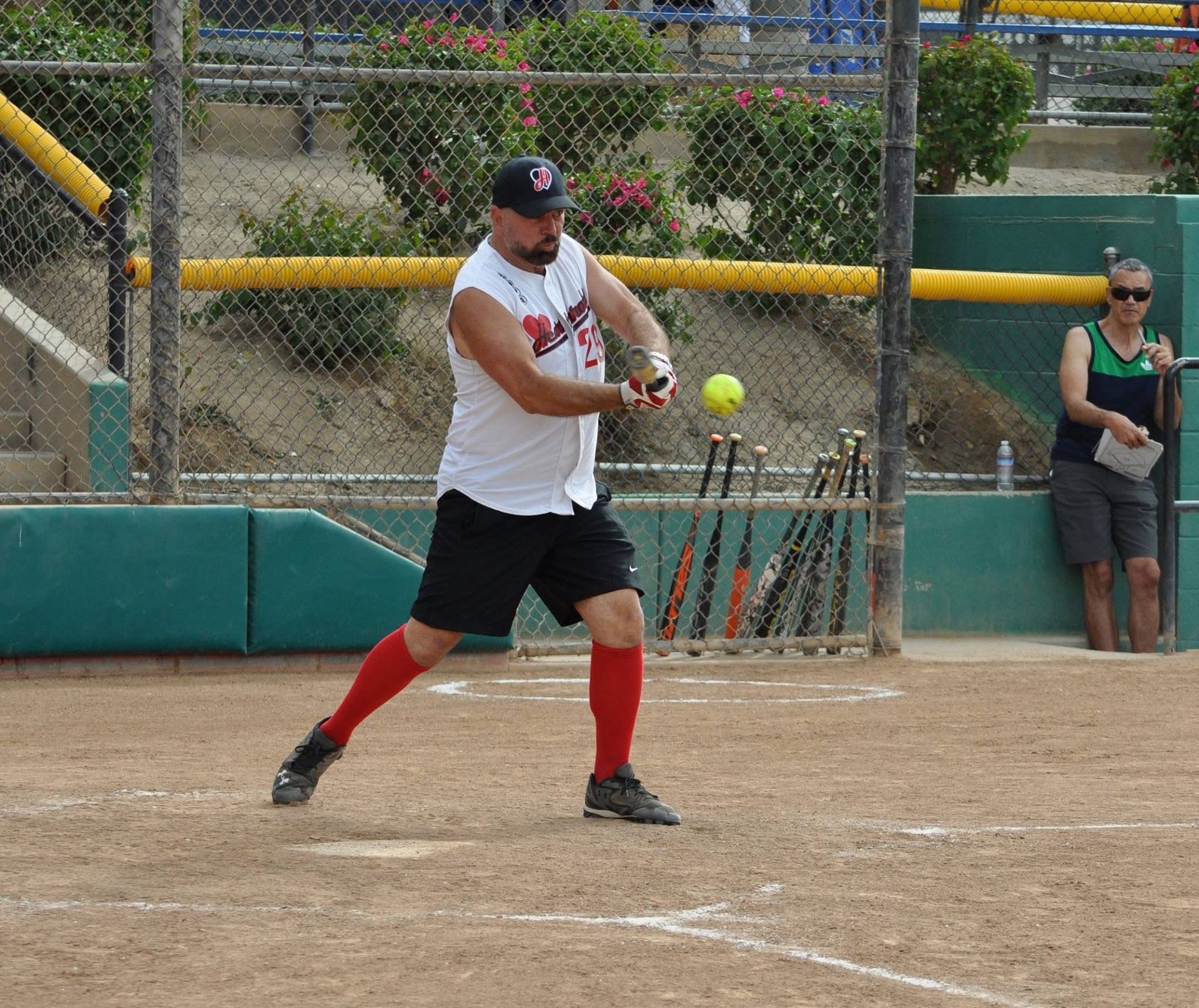 Softball batter in white shirt and black shorts swings at a yellow ball on a dirt field.