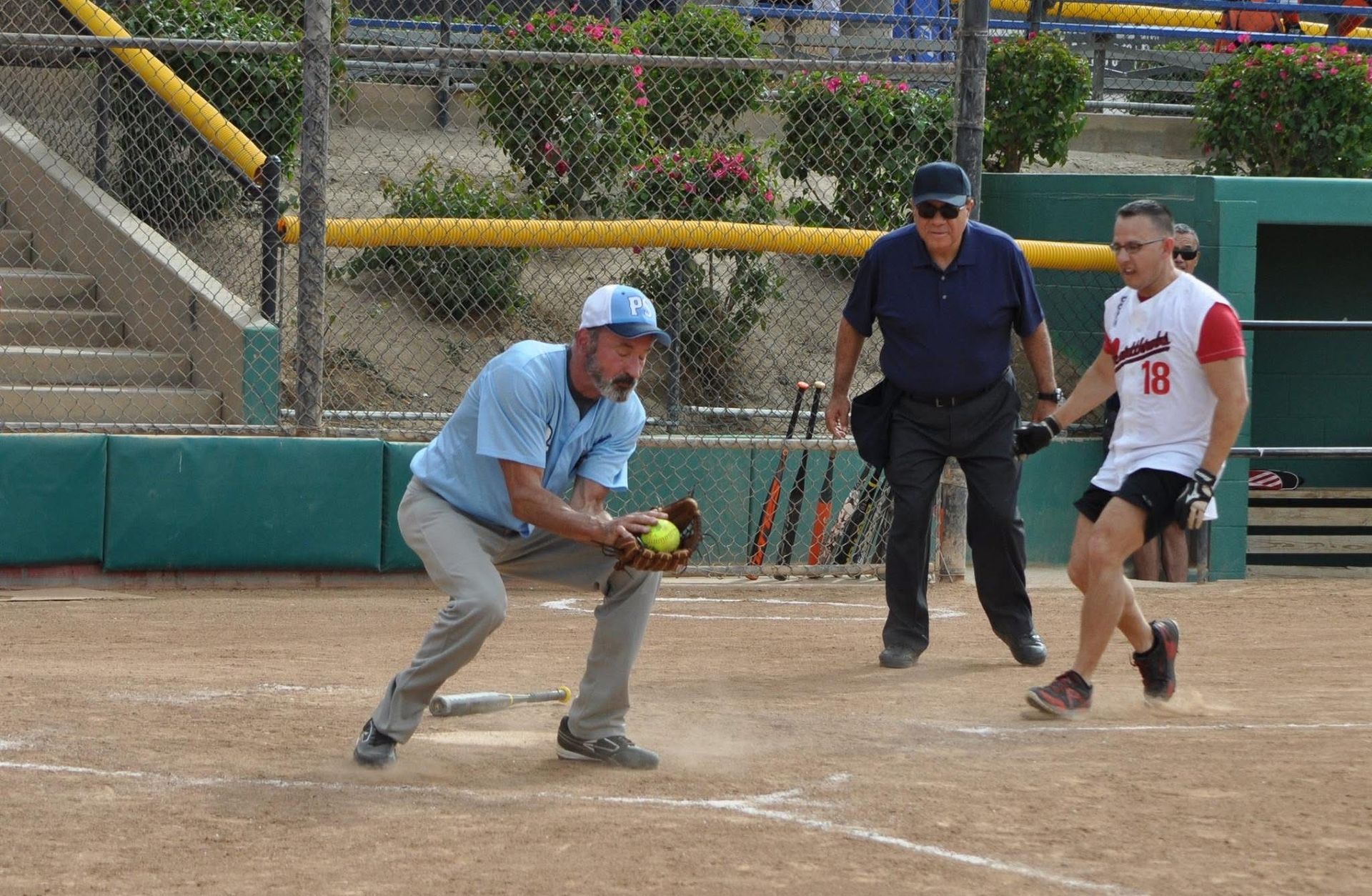Softball player in blue fielding a ground ball as batter and umpire watch at home plate