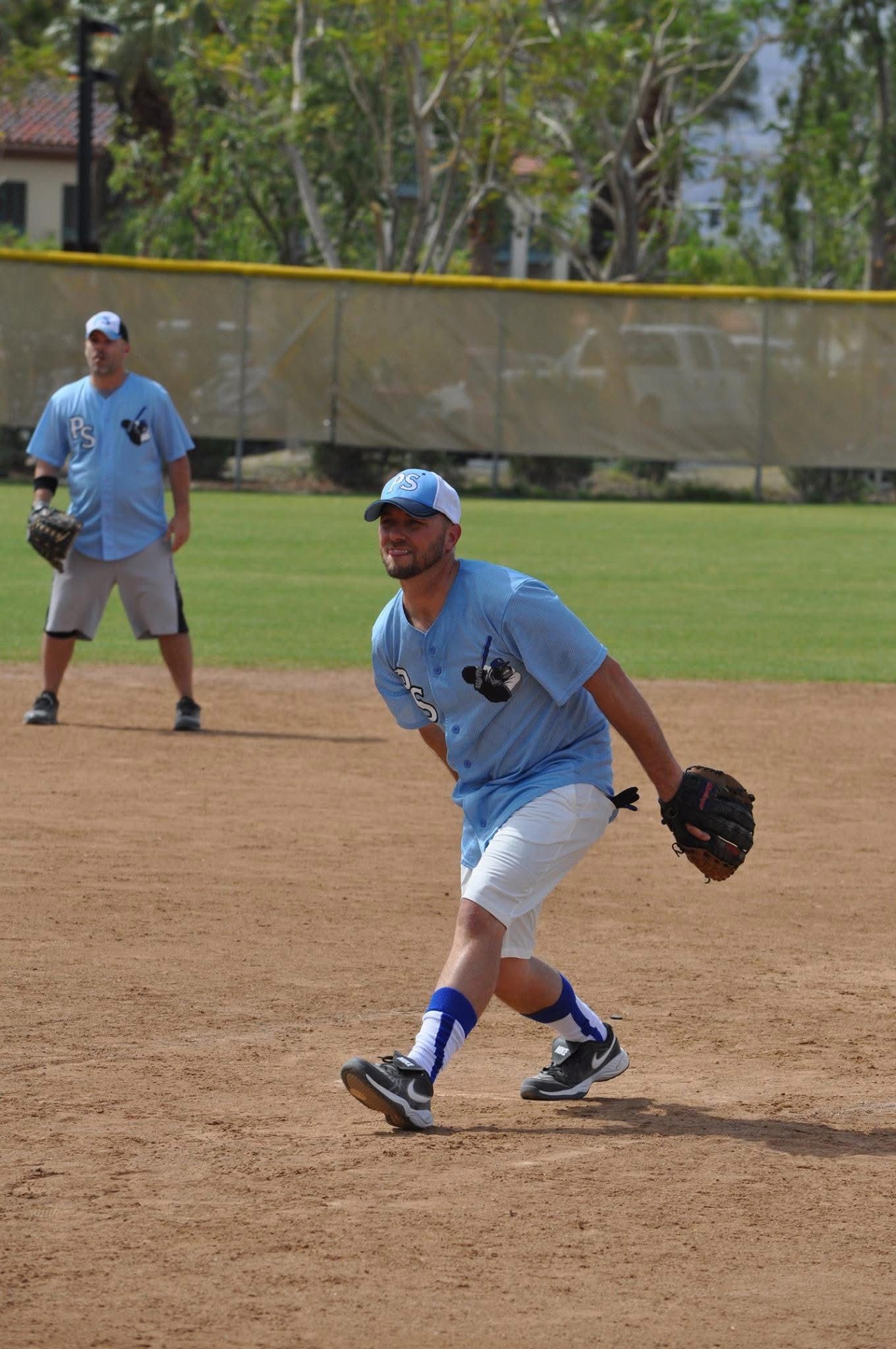 Baseball infielder in blue uniform fielding a ground ball on a dirt infield with a teammate in the background