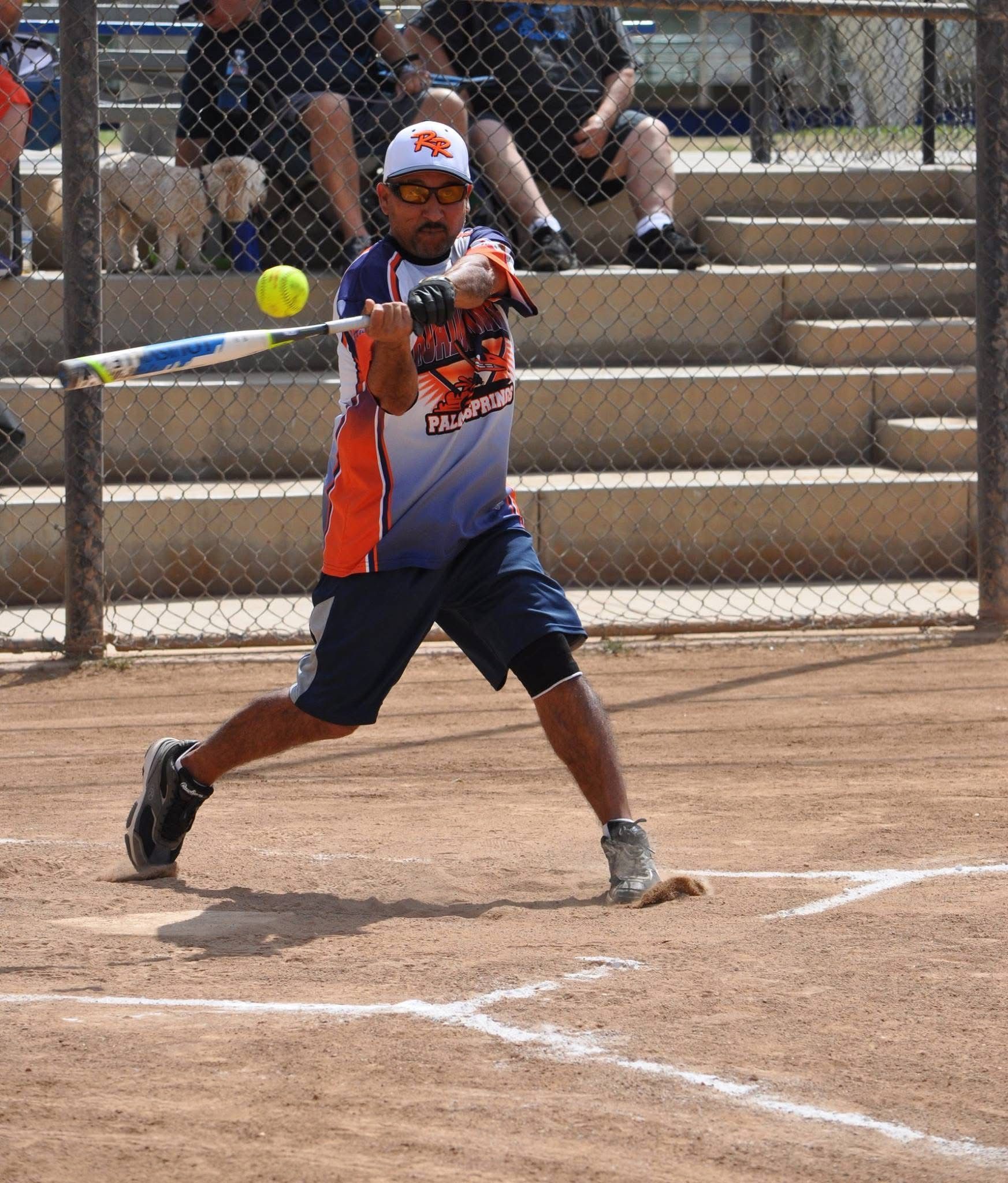 Softball player in orange and white swings a bat at a pitch on a dusty field.
