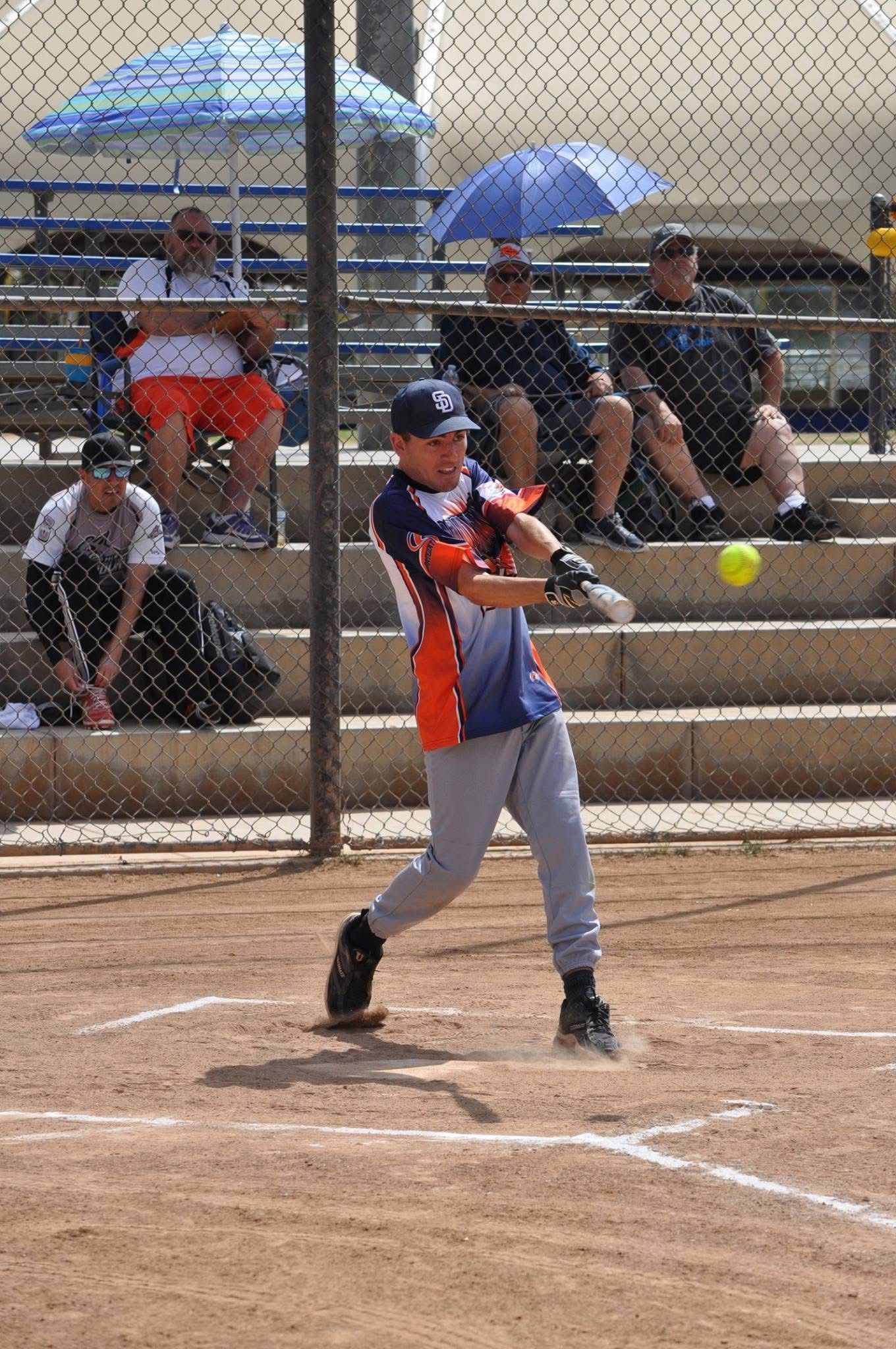 Youth softball batter in orange and navy swings at a pitch on a dirt field with spectators behind a fence