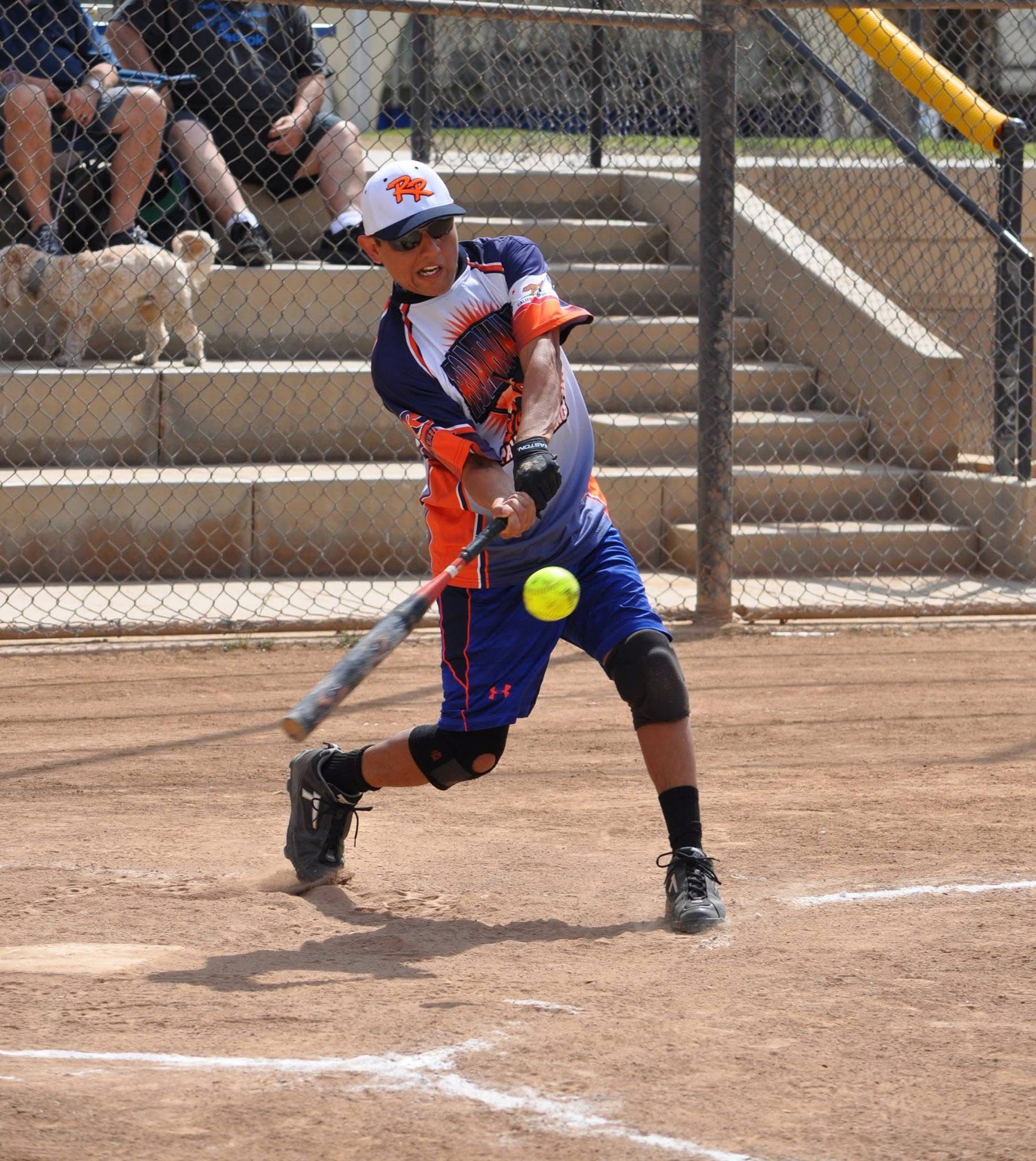 Softball player in orange and blue swings at a pitched ball on a dirt field.