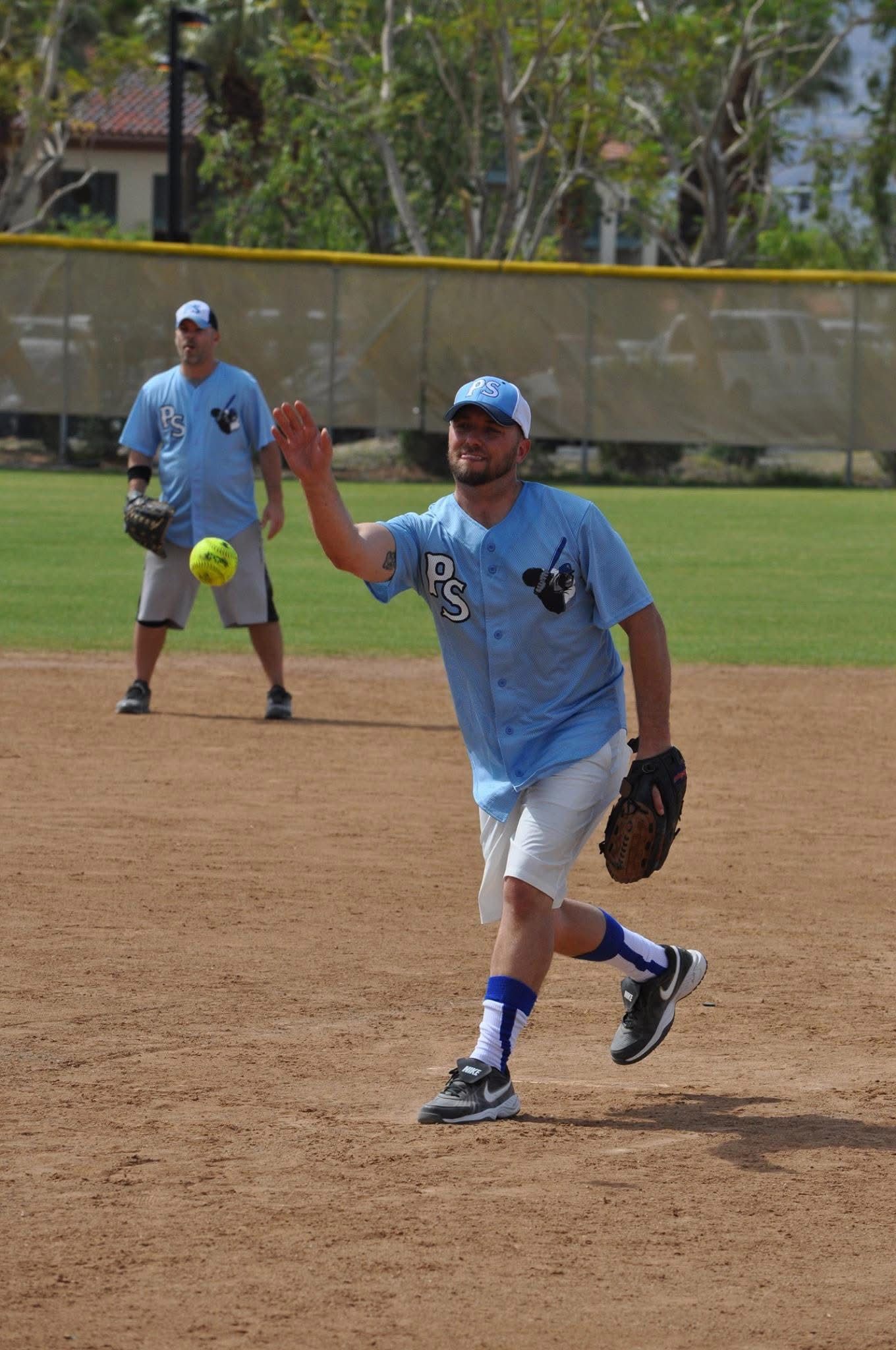 Softball player in blue fielding a ball on a dirt infield, with teammate in the background