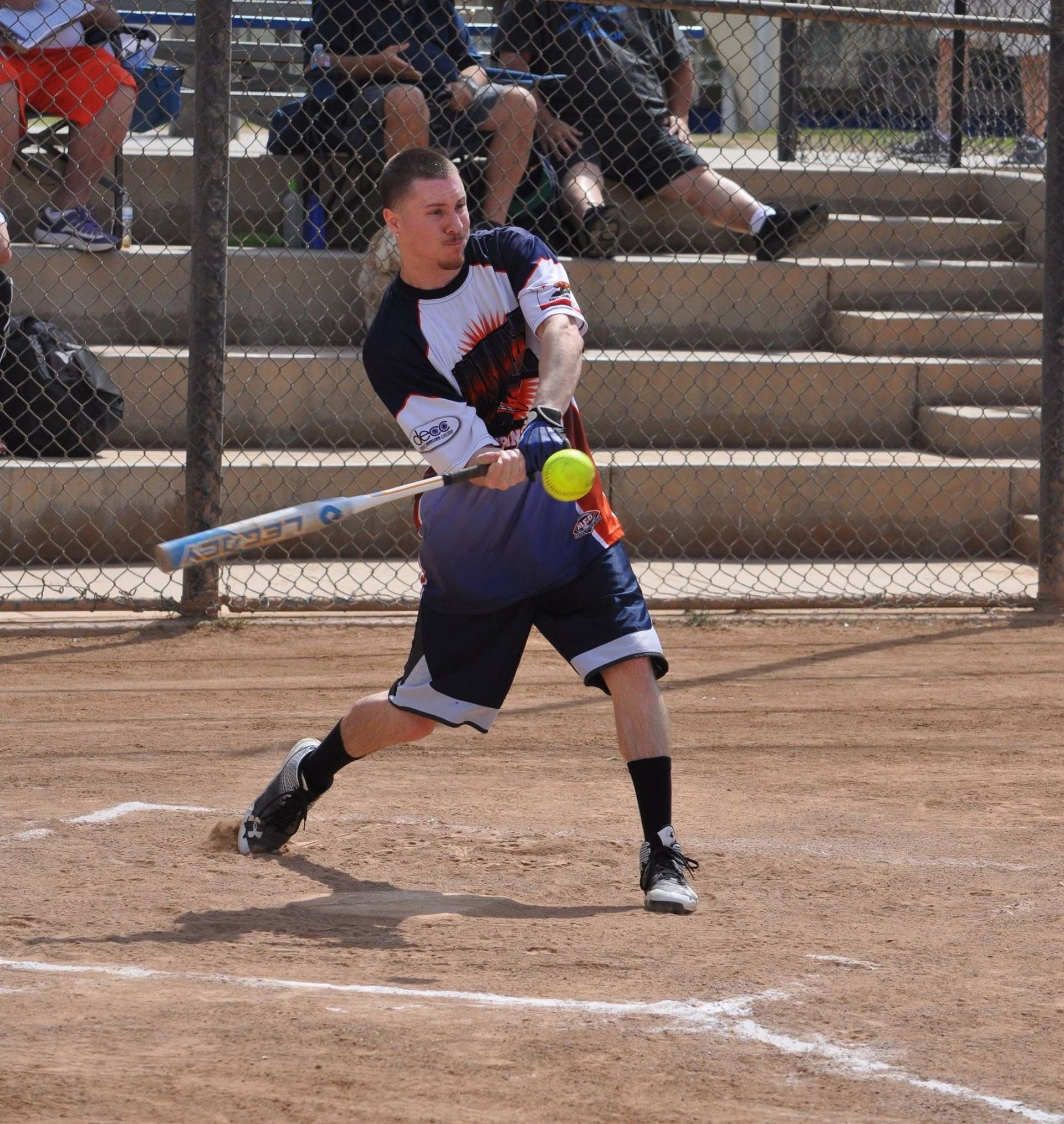 Softball player swings at a pitch on a dirt field, with a chain-link fence and bleachers behind.