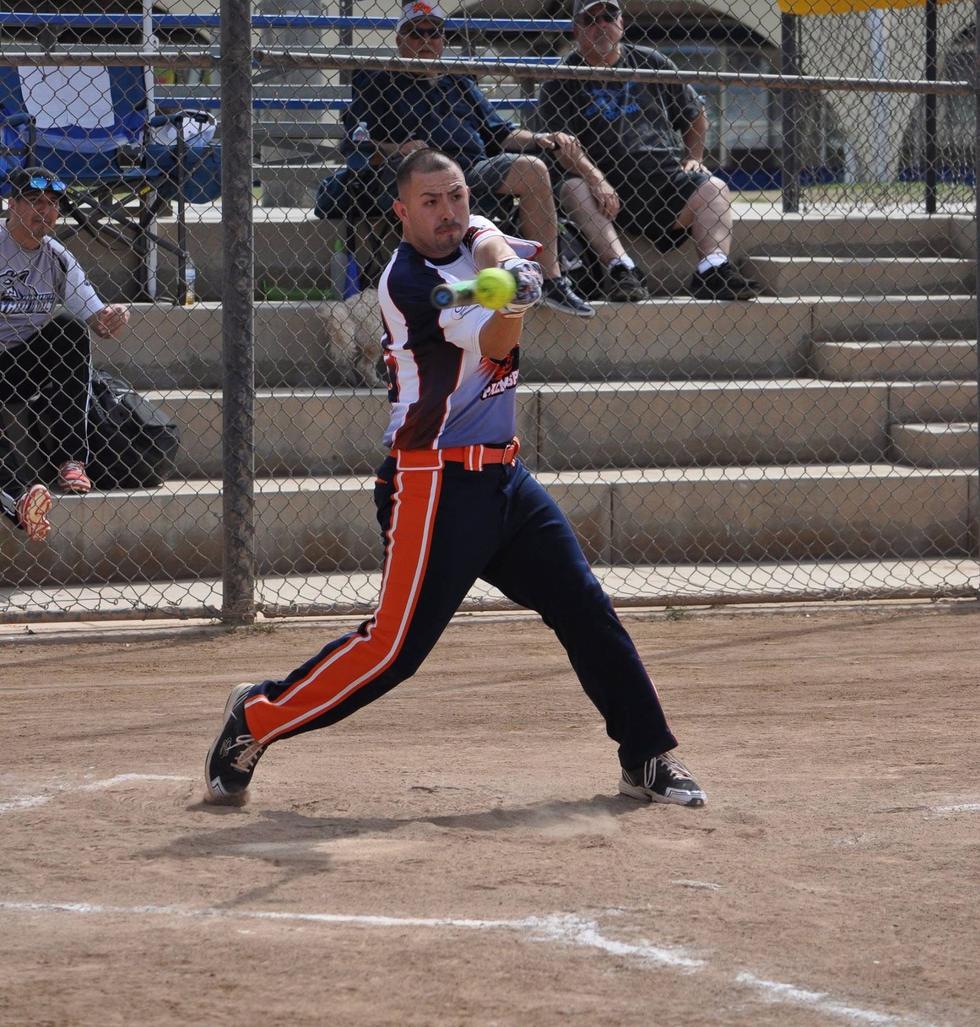 Softball player in navy and orange swings at a pitch on a dirt field, with bleachers and chain-link fence behind.