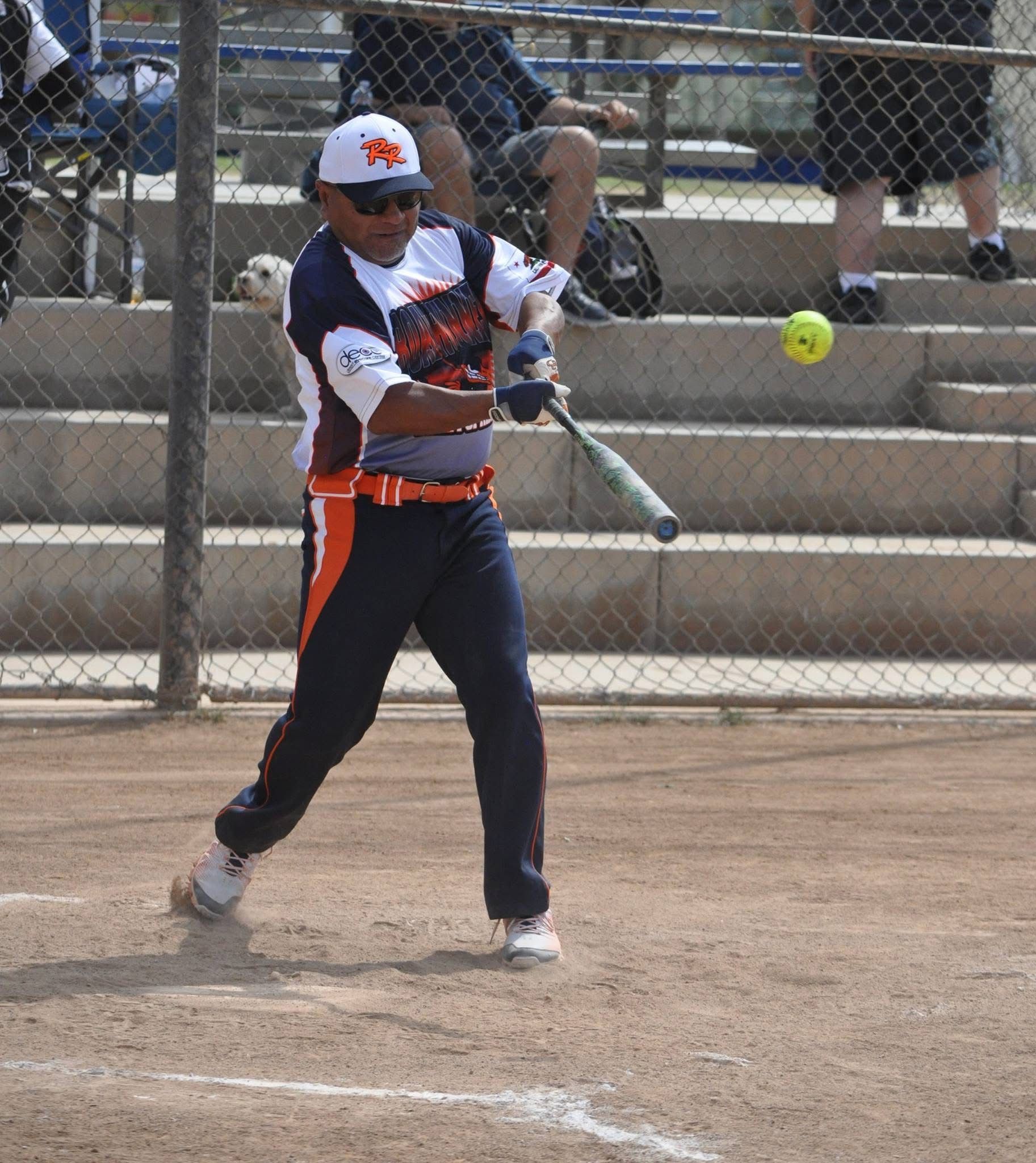 Softball player in black and orange uniform swinging a bat at a pitch on a dirt field