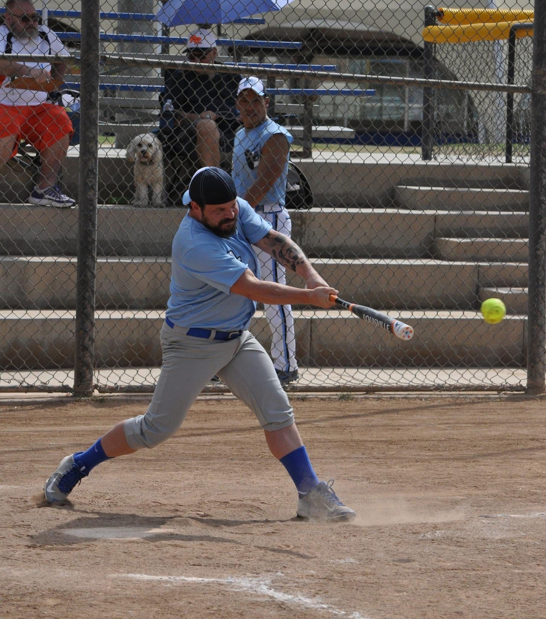Softball player in blue swings at a pitch on a dirt field, with the ball just ahead of the bat.
