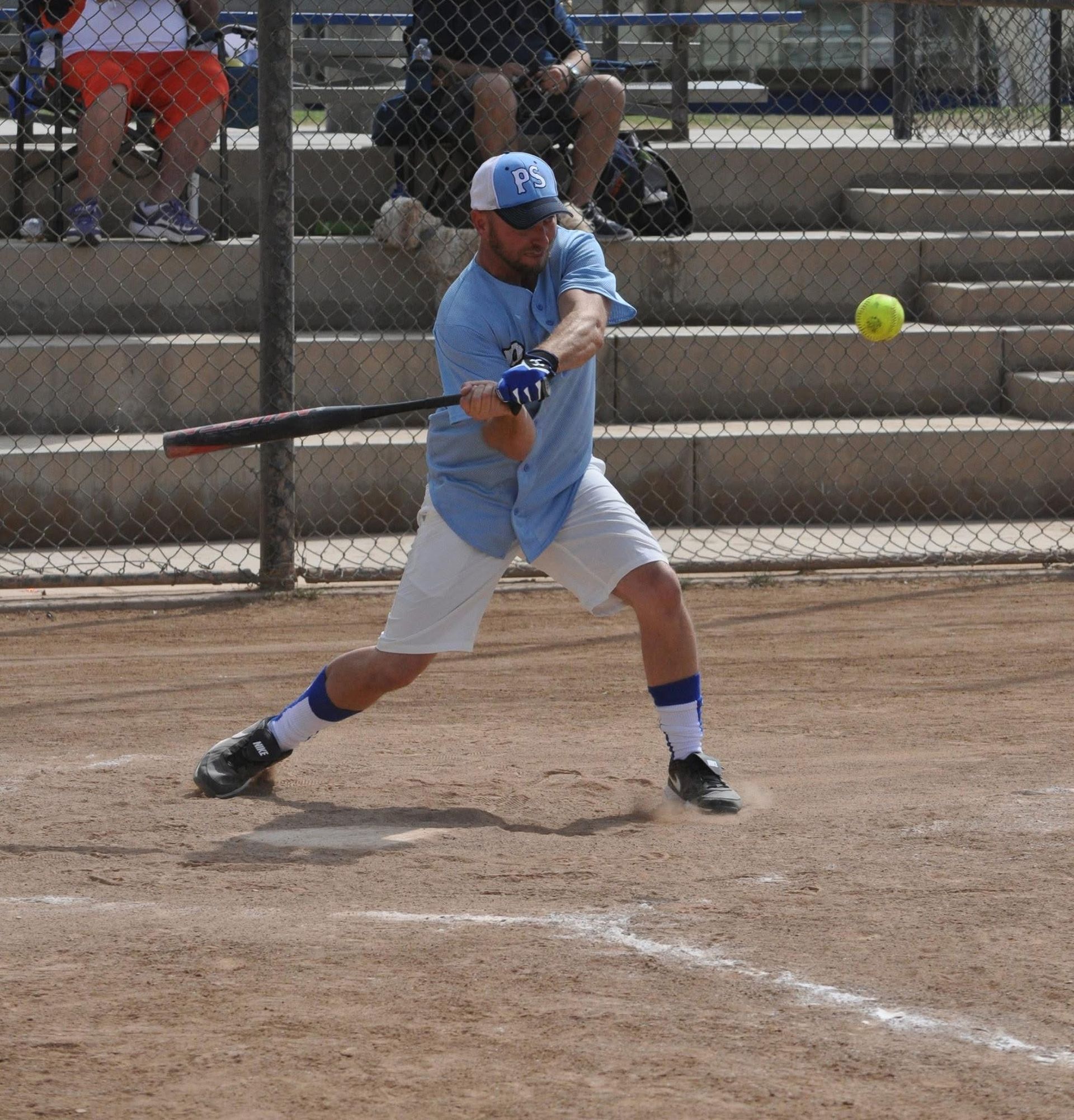 Softball player in blue swings at a pitch on a dirt field near bleachers.