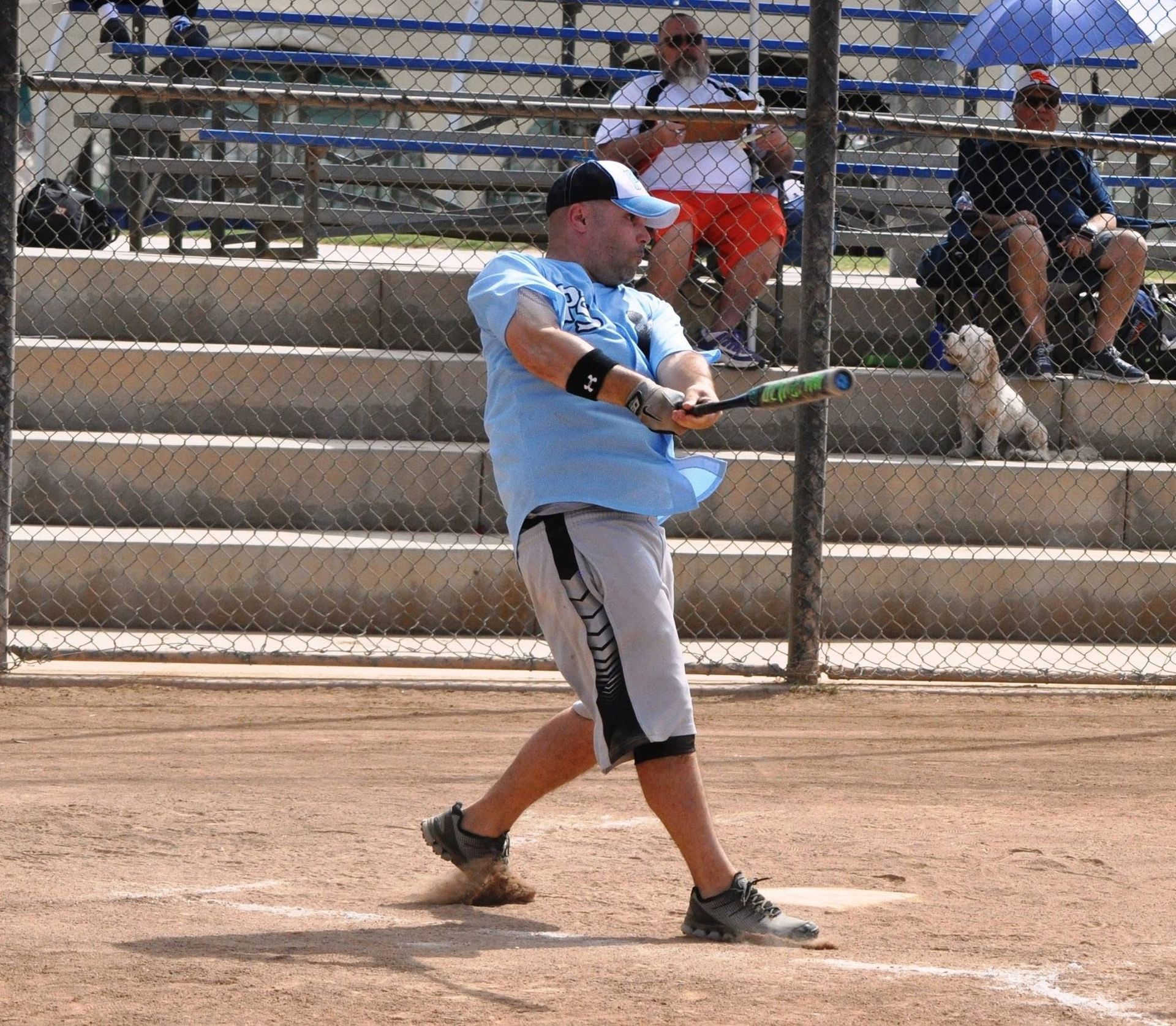 Baseball player in light blue swings a bat on a dirt field with bleachers in the background