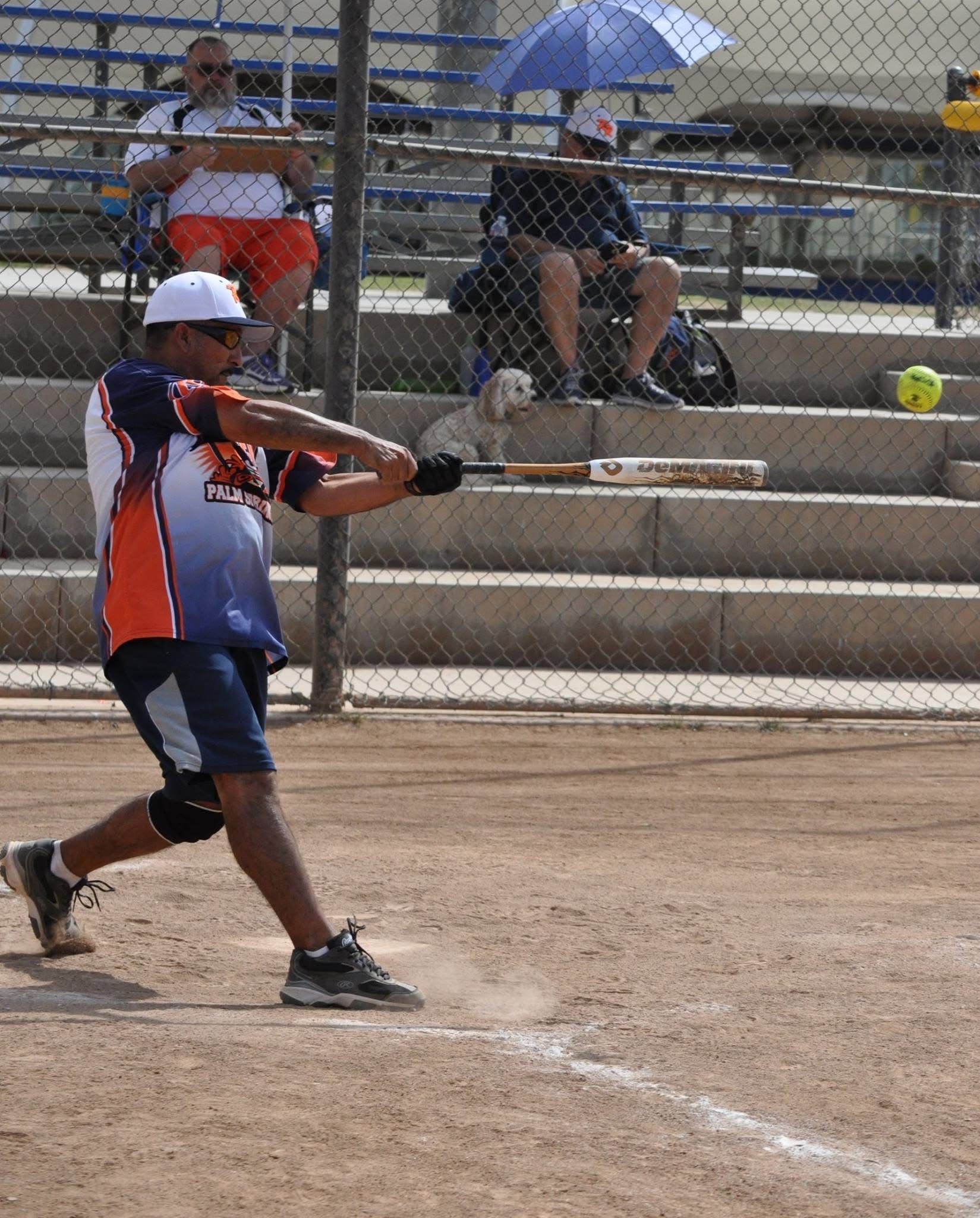 Softball player in blue and orange swings a bat on a dirt field with spectators in bleachers behind a fence.