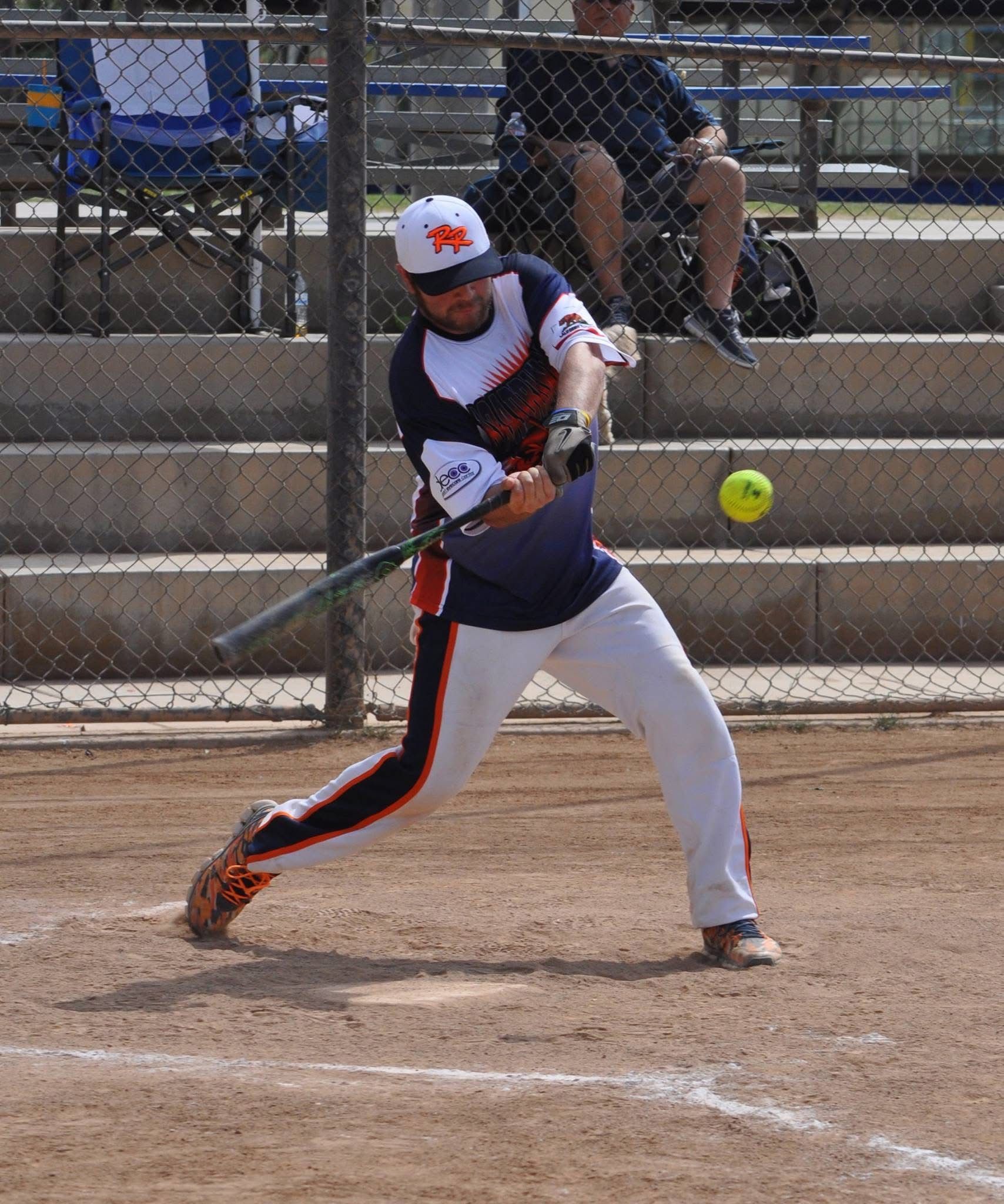 Softball player swings at a pitch on a dirt field, with a chain-link fence and bleachers behind.