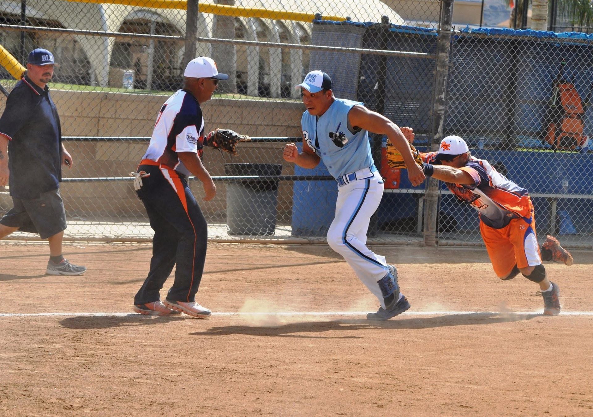 Softball players sprinting around a base during a dusty game, with fielders nearby.