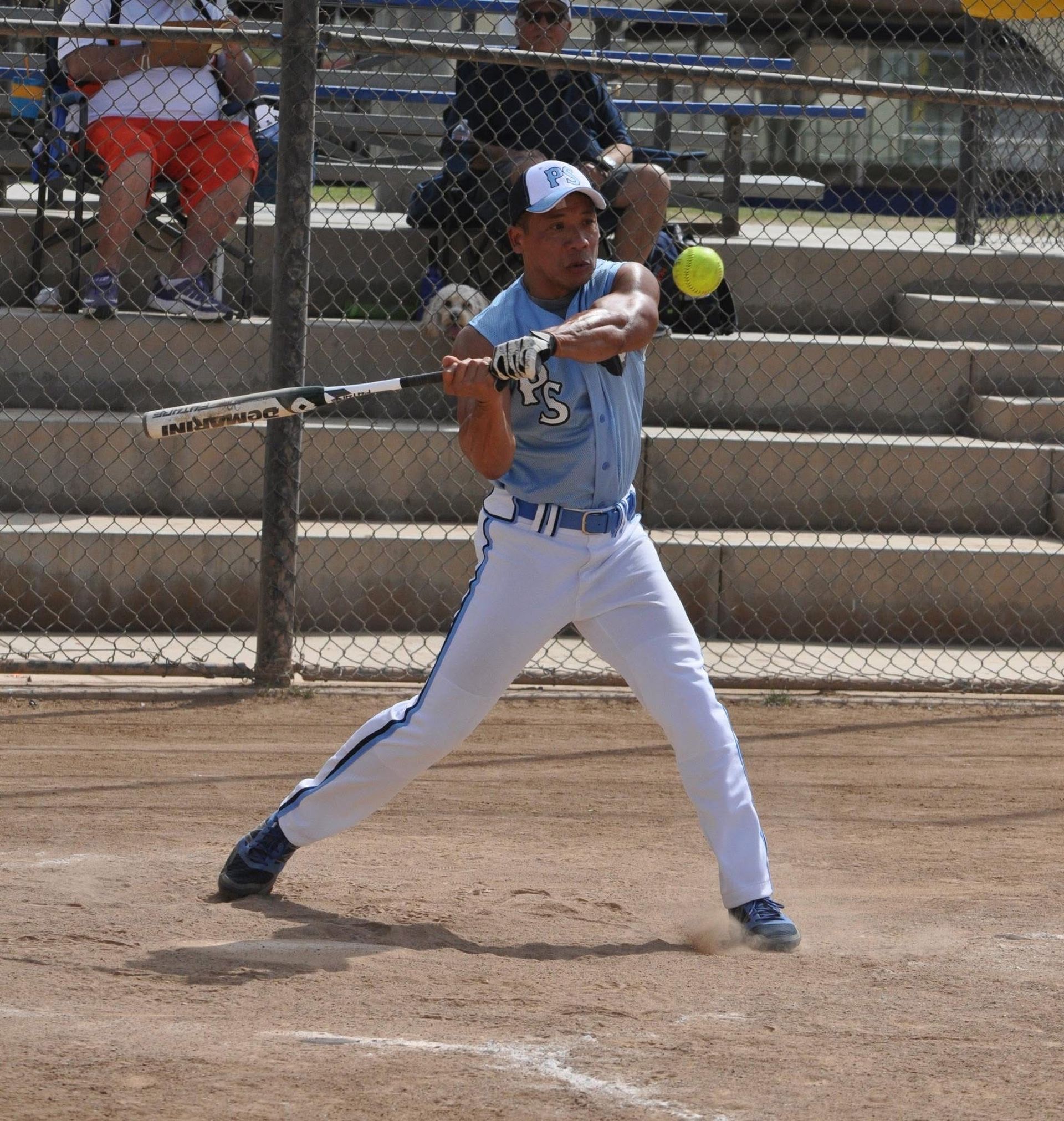 Softball player in white and blue uniform mid-swing at bat on a dirt field.
