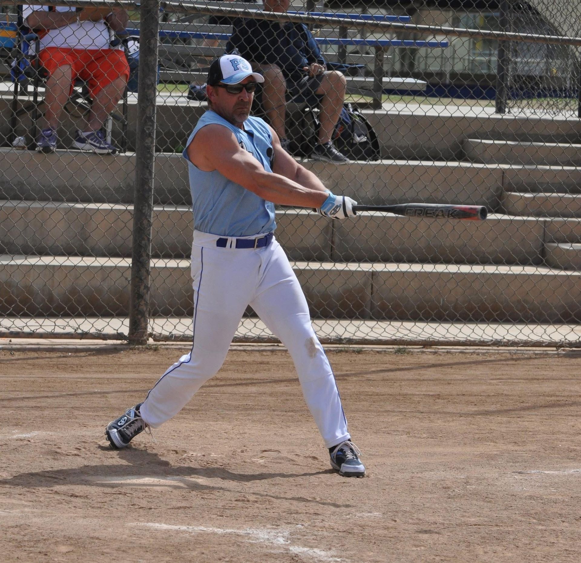 Baseball player in a light blue jersey and white pants swings a bat on a dirt field.