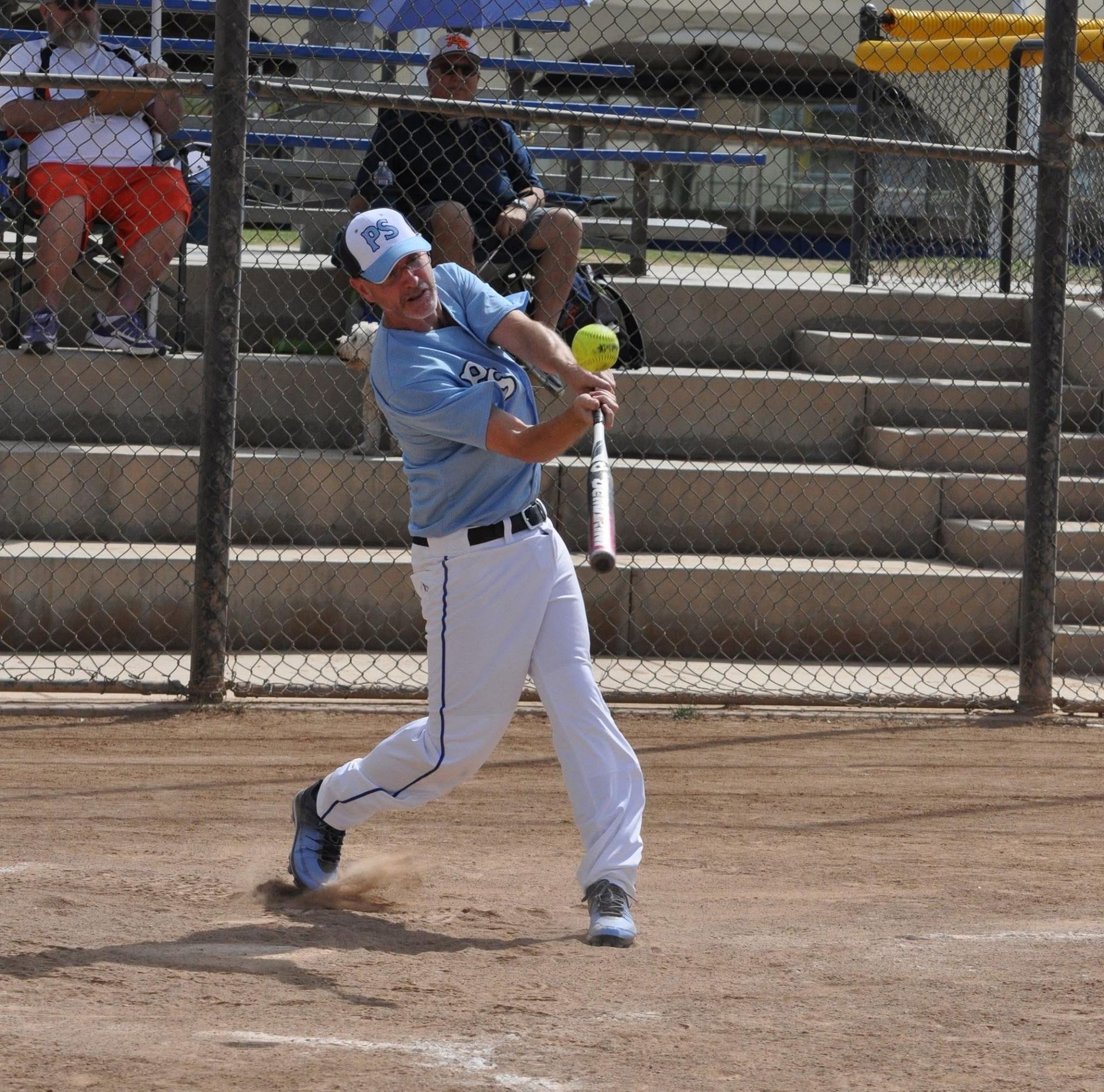 Softball player in light blue jersey swinging a bat on a dirt field with bleachers behind him