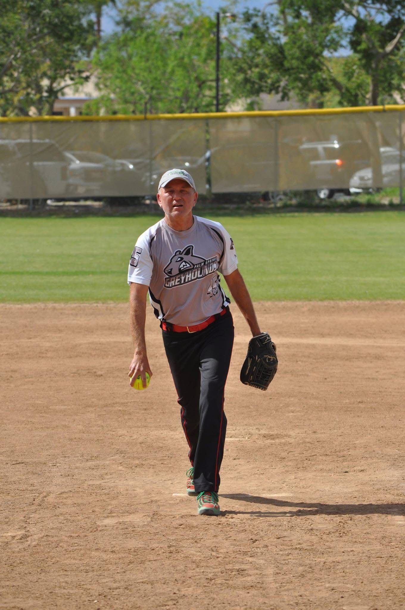 Softball player in white jersey and black pants walking on the infield, holding a glove and ball