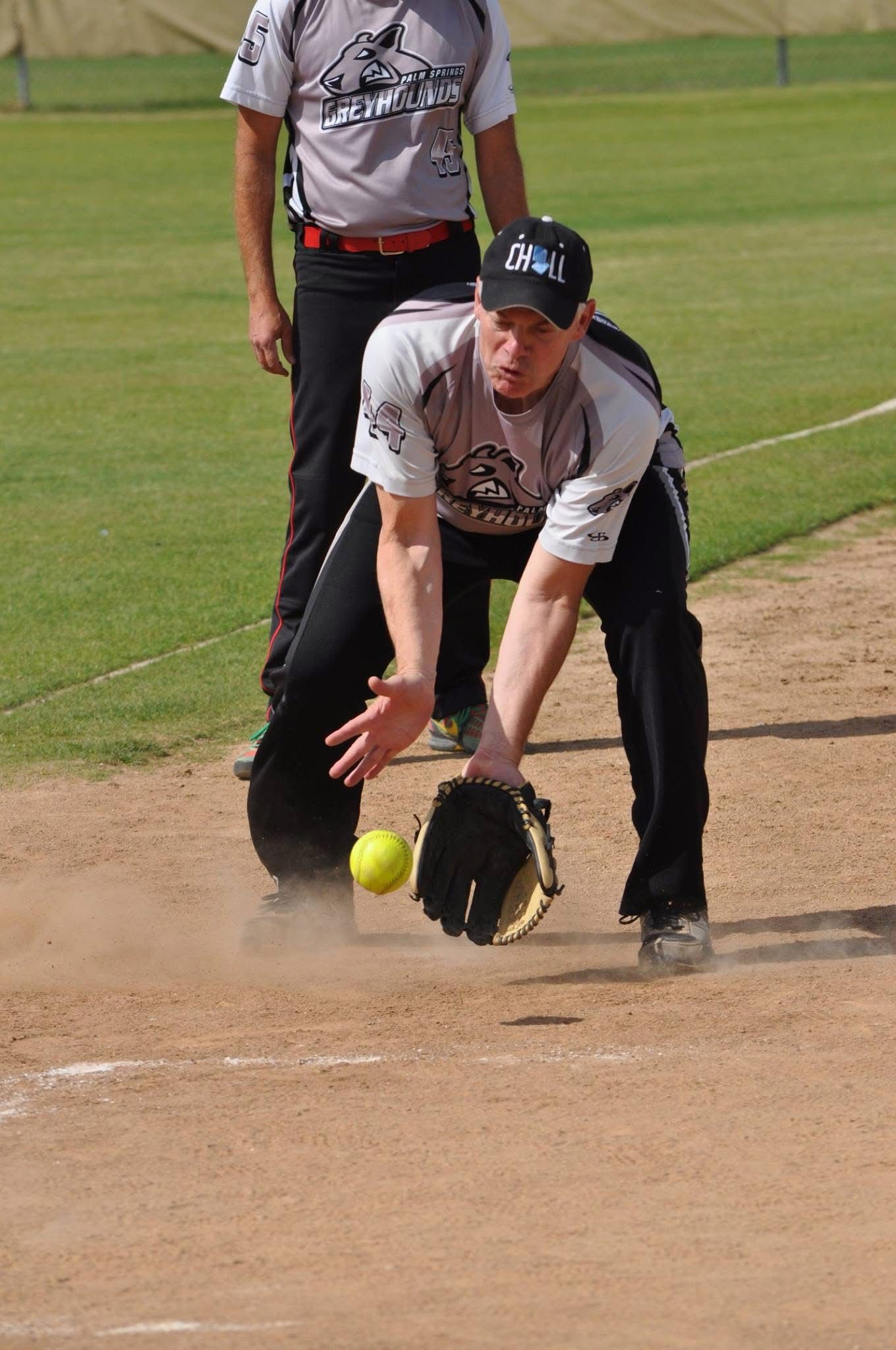 Softball player fielding a ground ball on a dirt infield, with a teammate behind him.