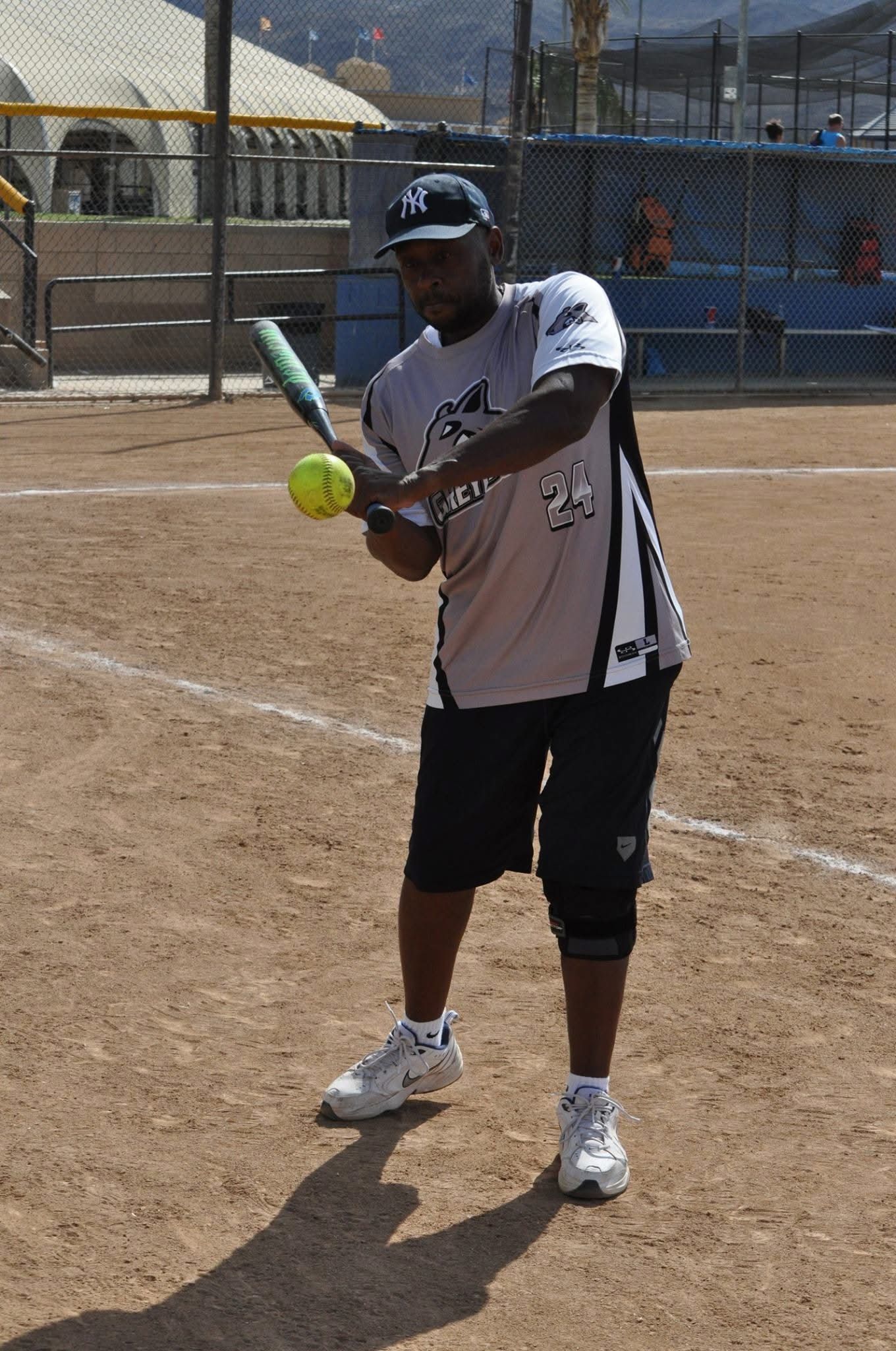 Softball player at bat on a dirt field, holding a green bat and yellow ball, with a chain-link backstop behind him