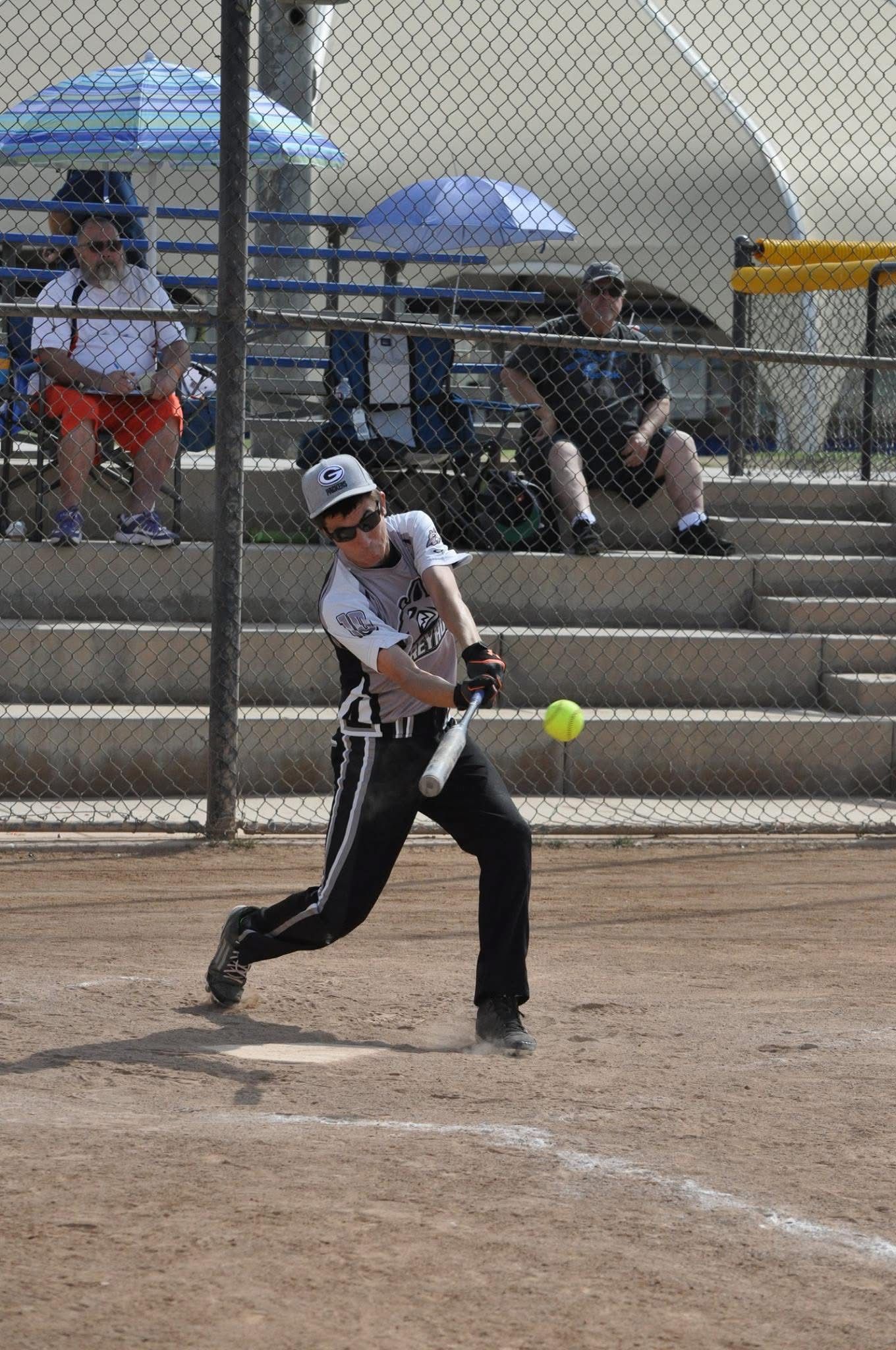 Softball batter in white and black swings at a yellow ball on a dirt field, with bleachers behind her.