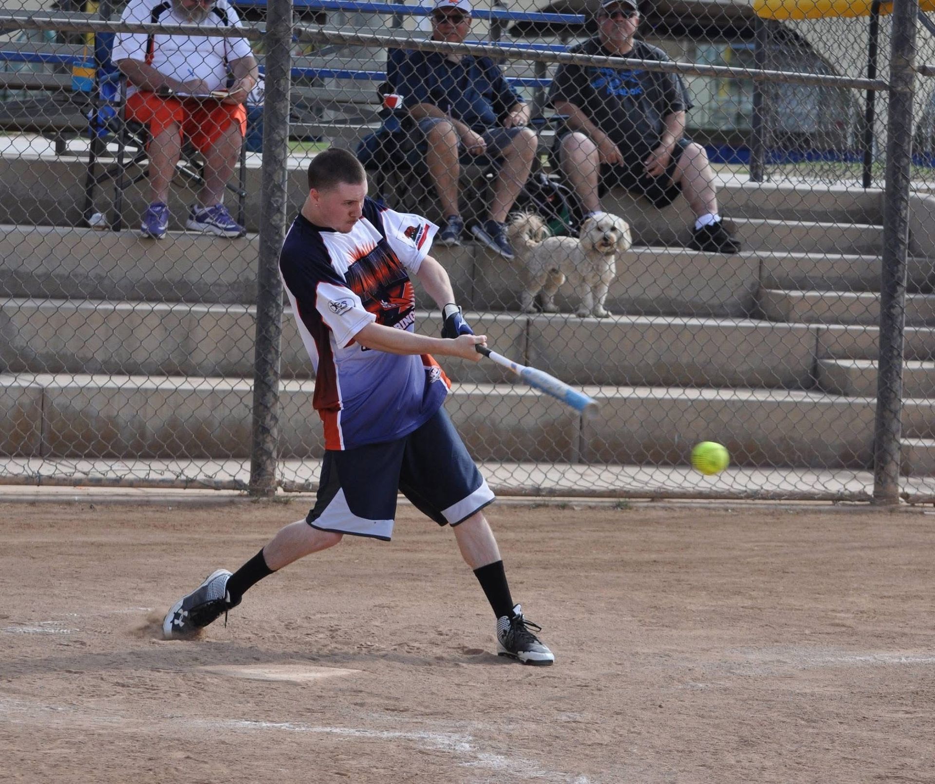Softball player in purple swings at a pitch on a dirt field, with spectators behind the fence.