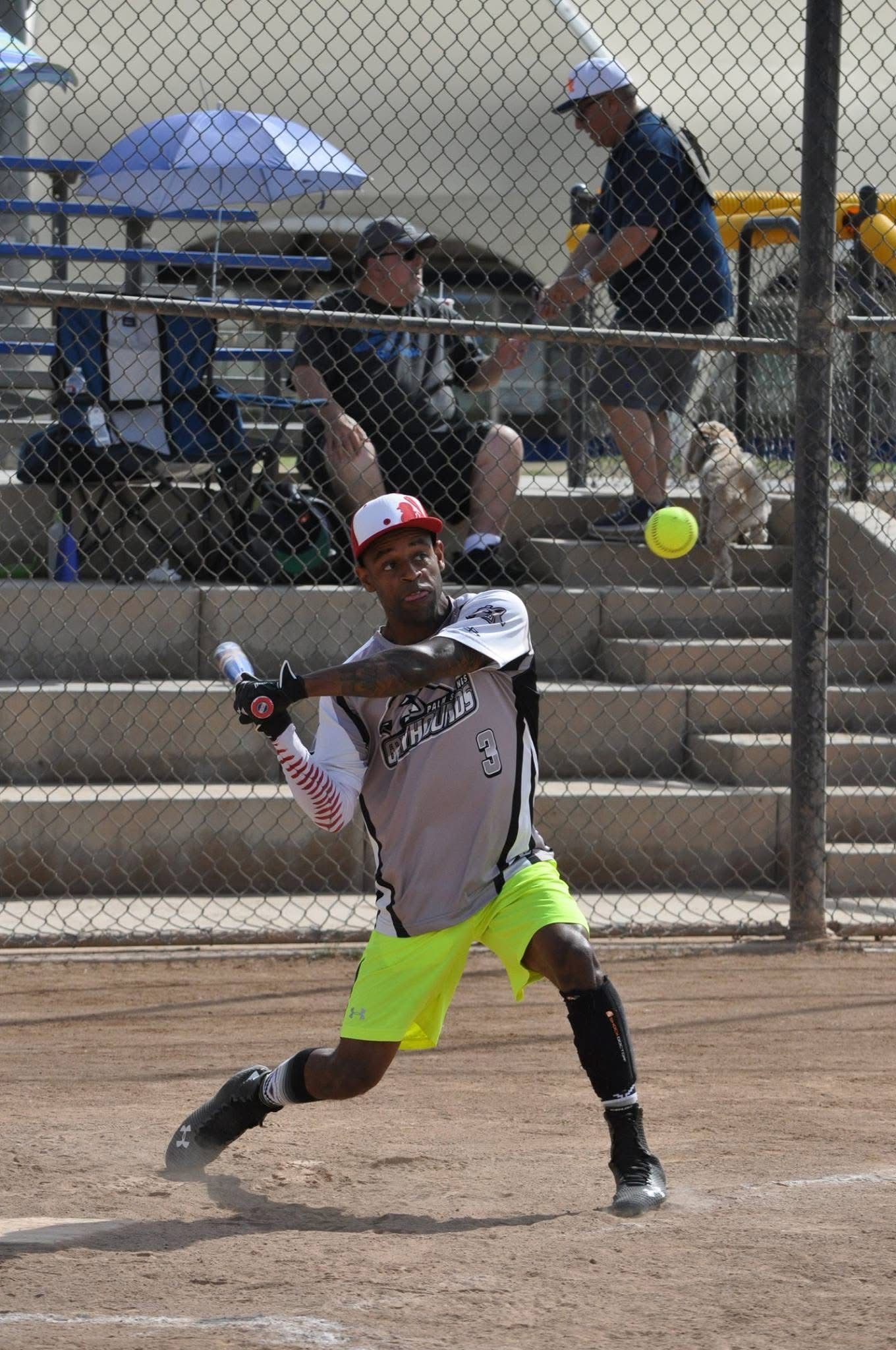 Baseball player in gray and neon shorts swings at a yellow ball on a dusty field, with bleachers behind him