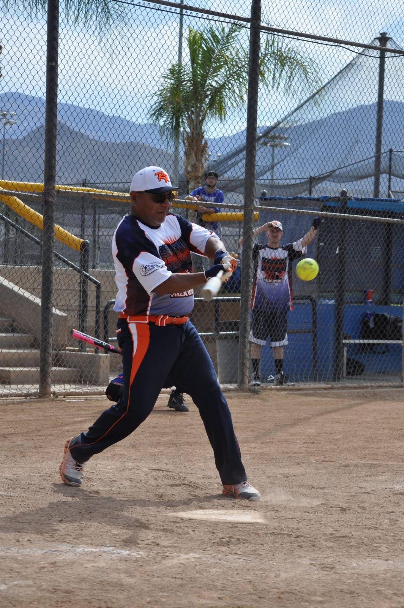 Softball player in white and navy swings at a pitch on a dirt field behind a netted fence.