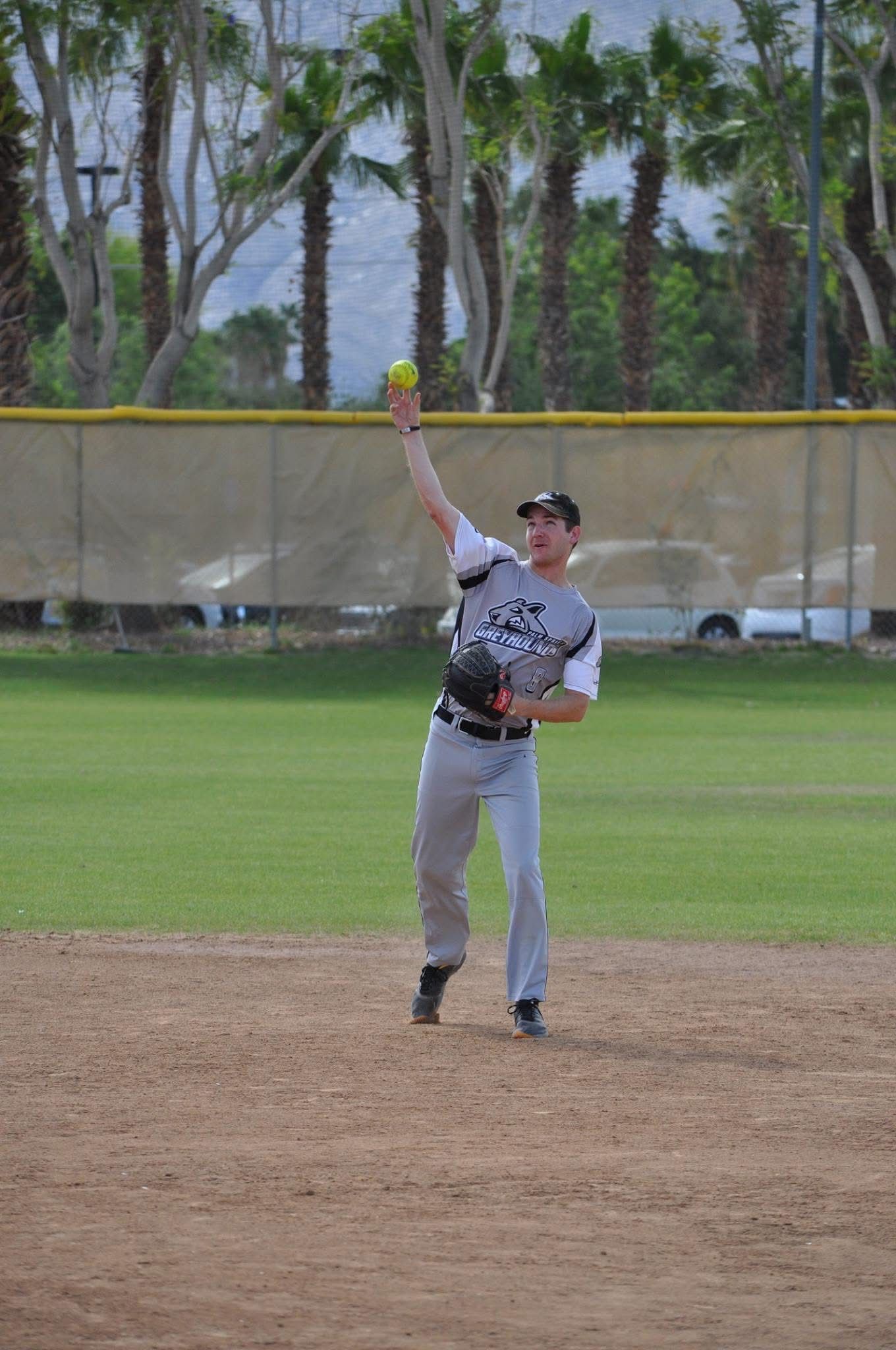 Baseball player in gray uniform reaching high to catch a ball on a dirt infield, with outfield fence and palm trees behind.